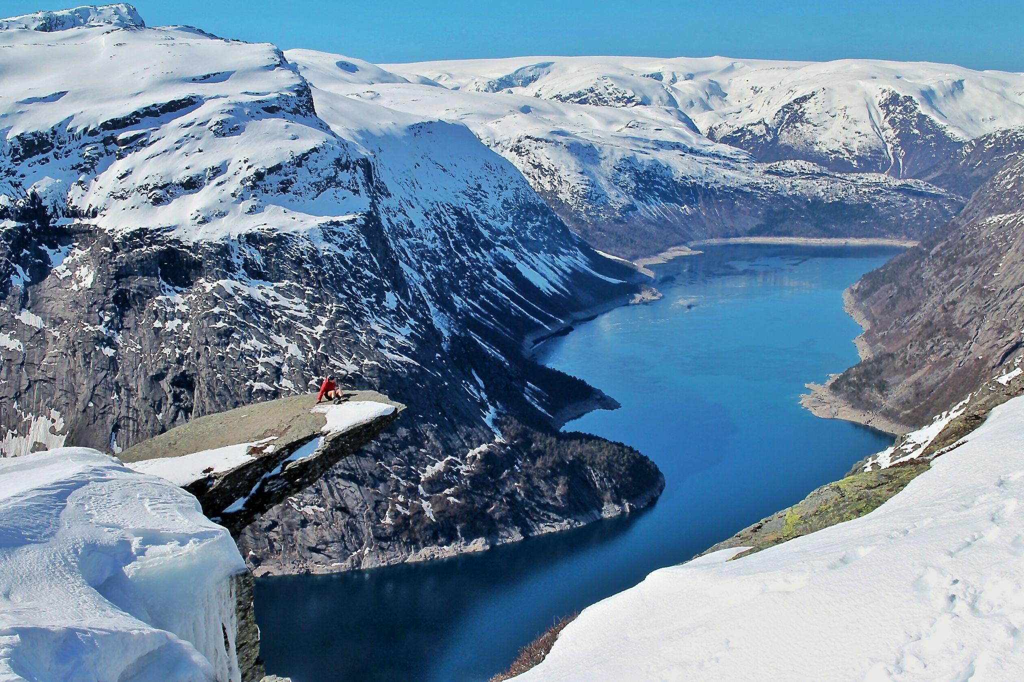 Person sit på Trolltunga om vinteren med utsikt over den snødekte fjorden og fjellandskapet.