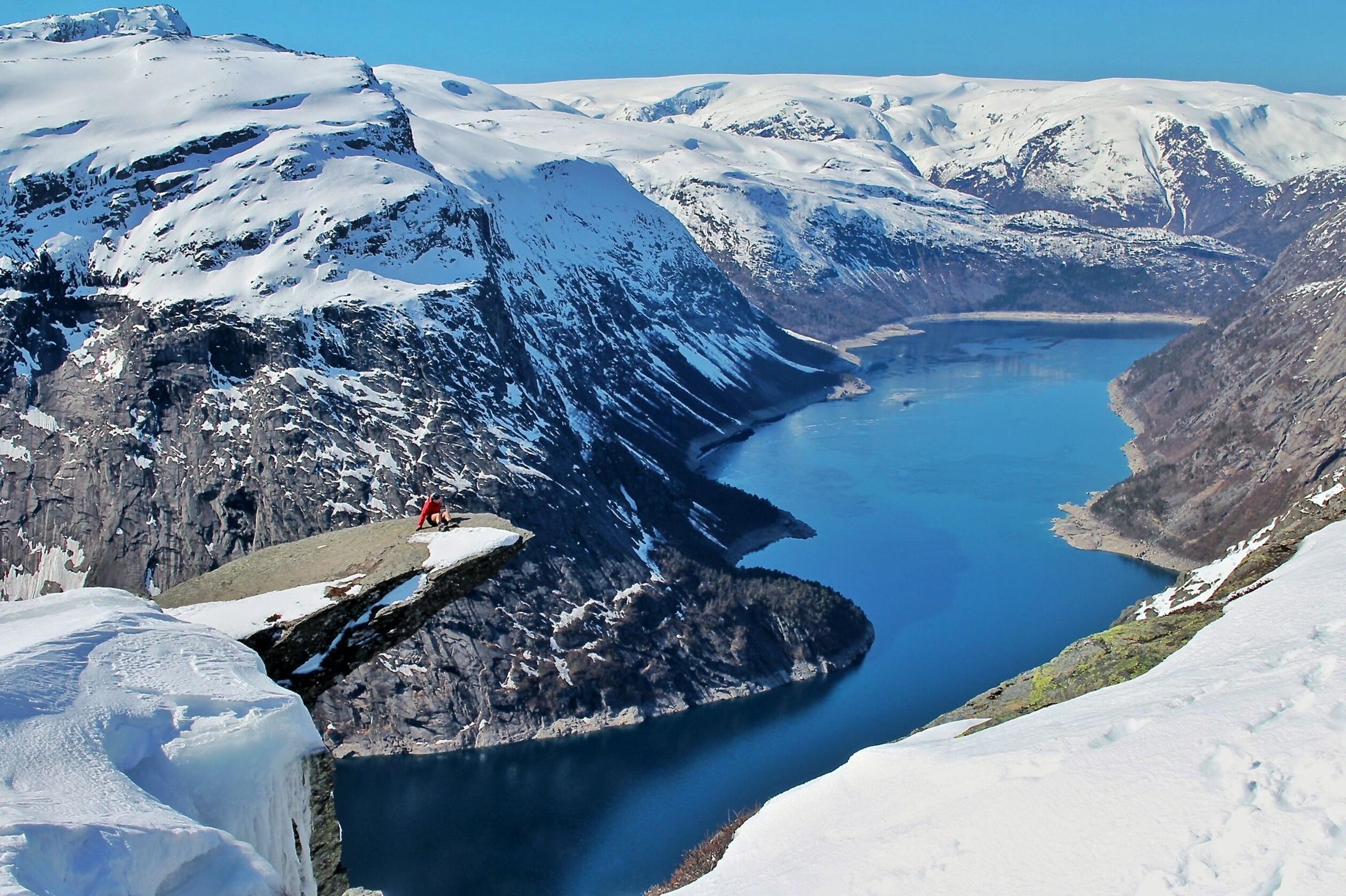 Person sit på Trolltunga om vinteren med utsikt over den snødekte fjorden og fjellandskapet.