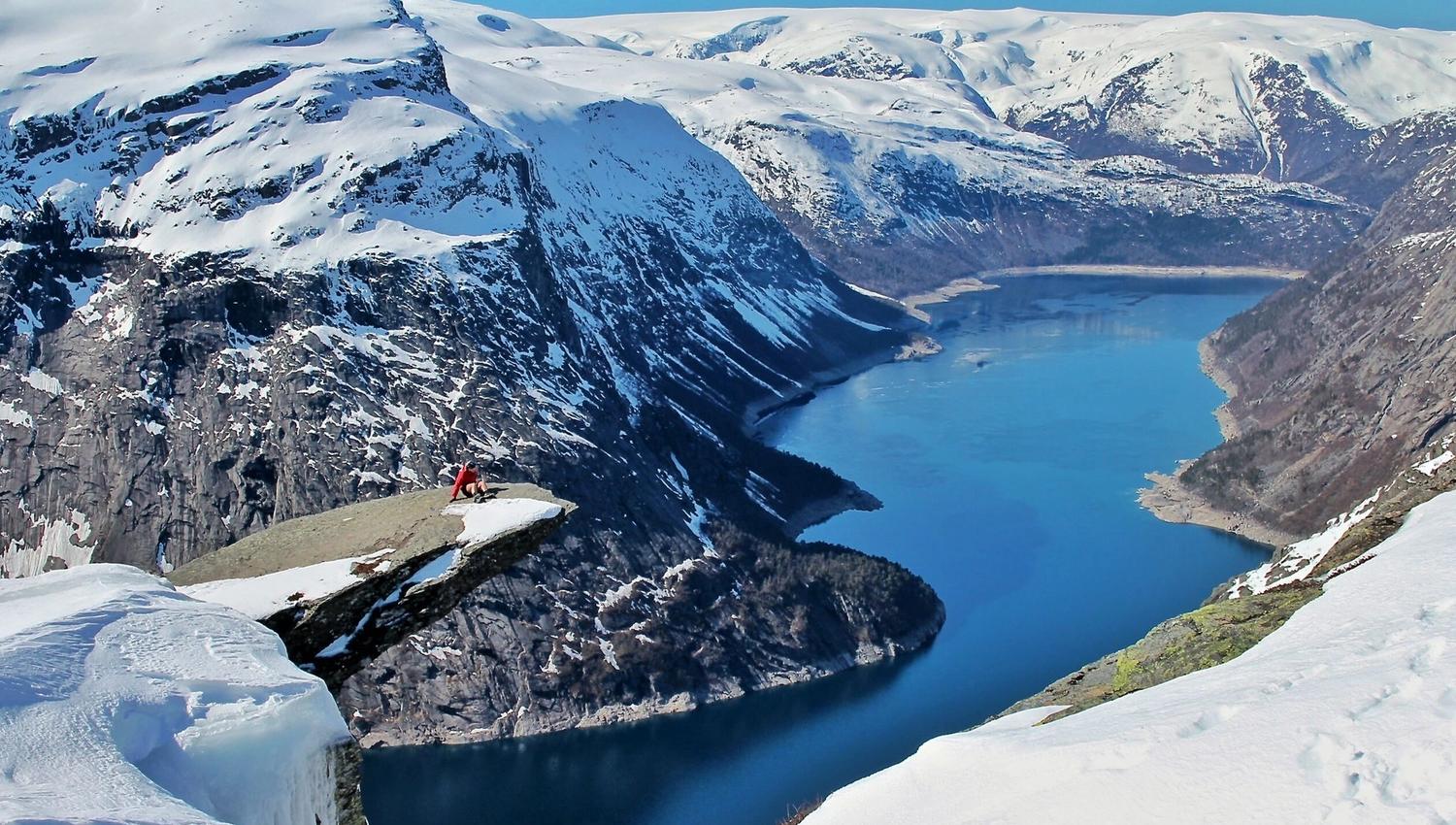 Person sitting on Trolltunga in winter with a view of the snow-covered fjord and mountains.