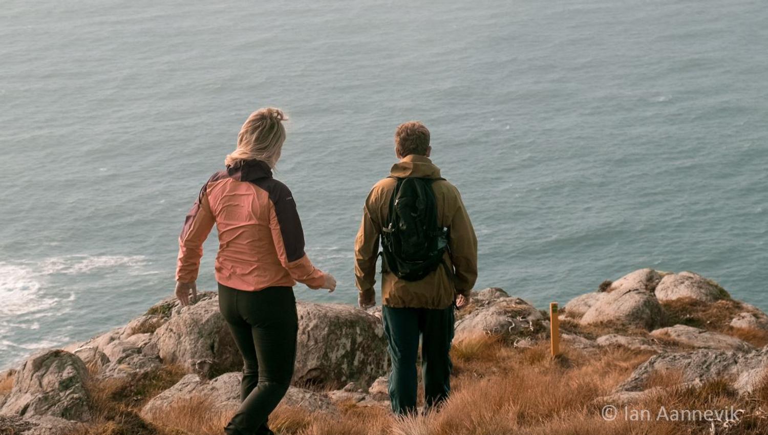 Two hikers in jackets and backpacks walk on a rocky coastal hillside, looking out over the calm ocean..