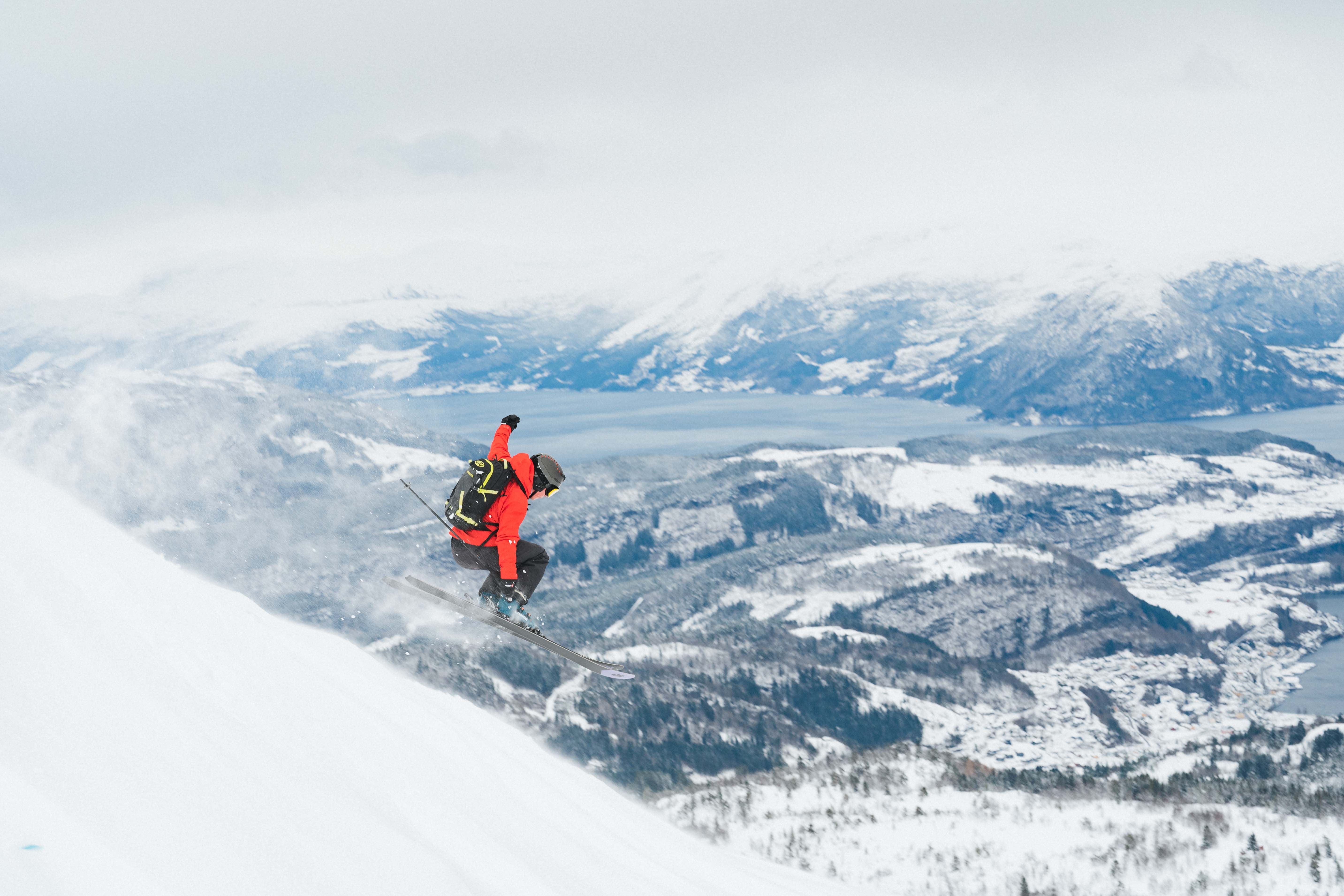 A person in a red jacket hops down a hill skiing. View towards a fjord and snow-covered landscape in the background. 