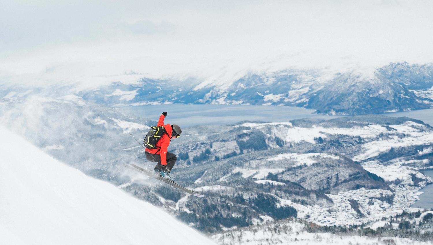 A person in a red jacket hops down a hill skiing. View towards a fjord and snow-covered landscape in the background.