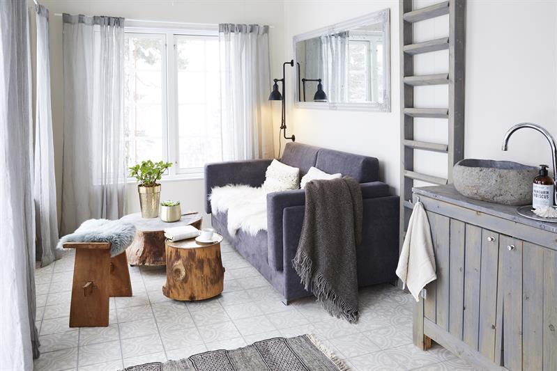 Lys og moderne stue med sofa, små trebord, kjøkkenkrok og store vinduer som slipper inn naturlig lys.