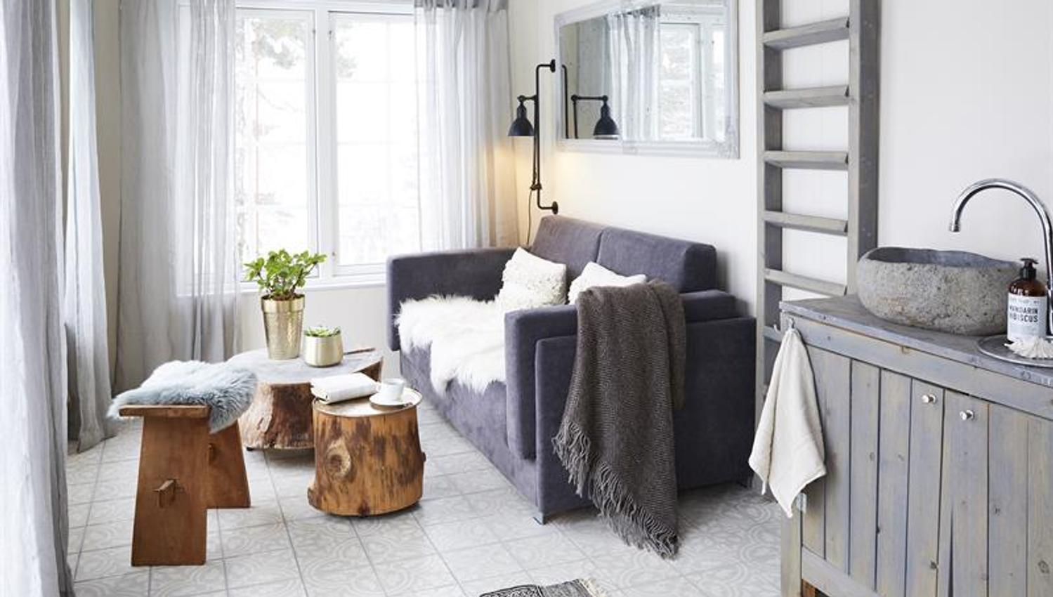 Lys og moderne stue med sofa, små trebord, kjøkkenkrok og store vinduer som slipper inn naturlig lys.