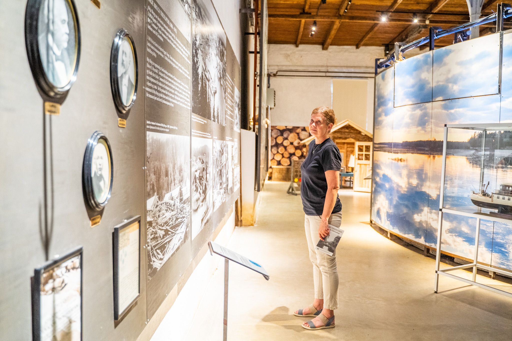 Bildet viser en kvinne som ser på en utstilling på et museum. Hun står foran en vegg med gamle bilder og tekst, og holder en vannflaske i hånden. I bakgrunnen sees en del av en utstilling med bilder av vann og tømmerstokker.