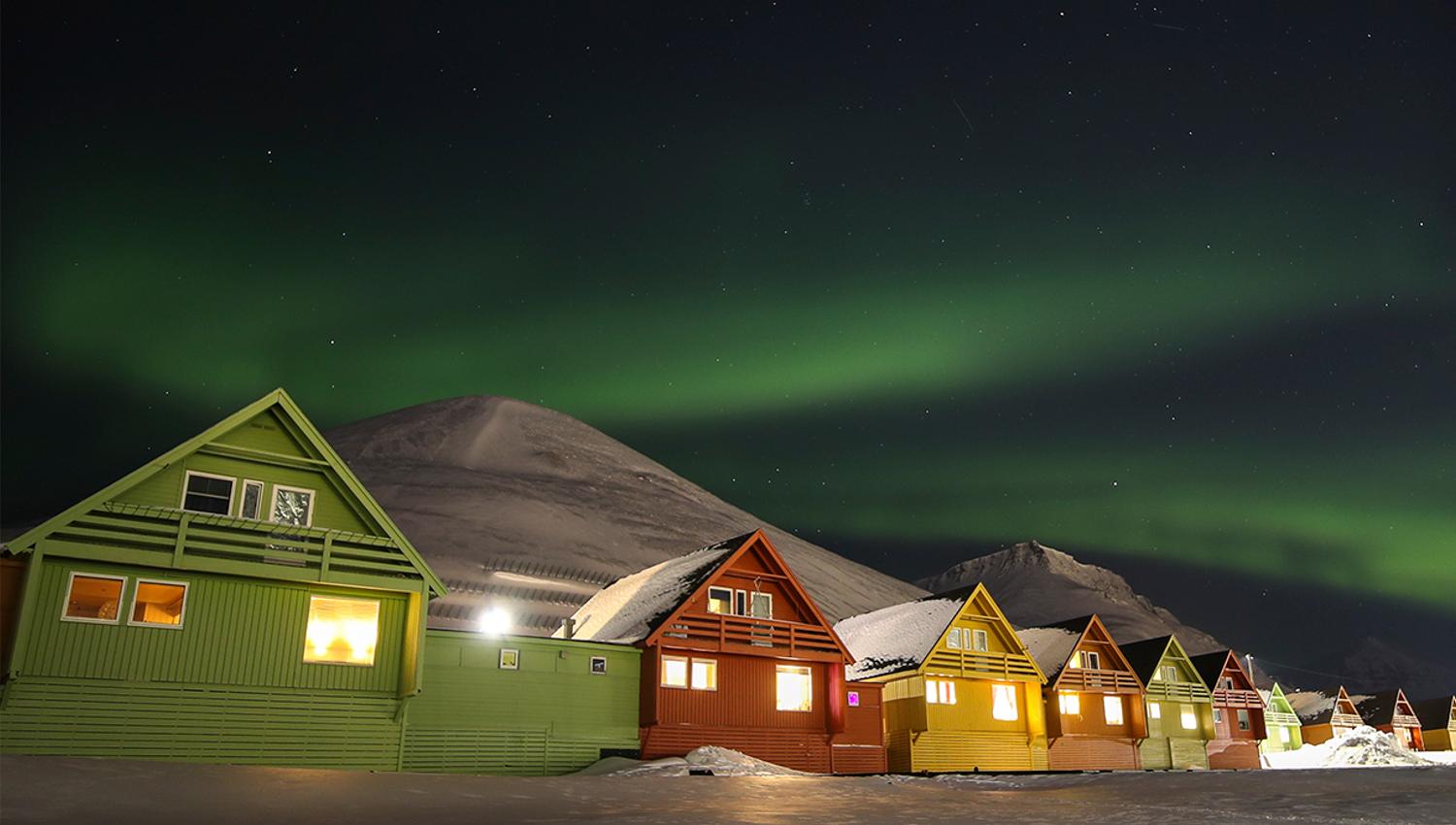 Fargerike hus i Longyearbyen med snødekte fjell og nordlys på himmelen i bakgrunnen