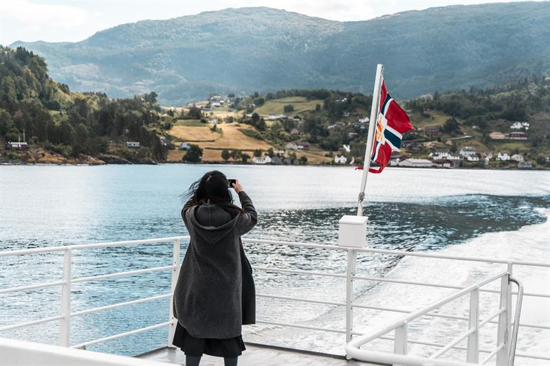 Passasjer fotograferer fjordlandskapet frå akterdekket under fjordcruise på Hardangerfjorden, norsk flagg i vinden.
