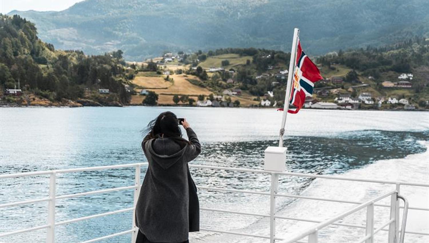 Passasjer fotograferer fjordlandskapet frå akterdekket under fjordcruise på Hardangerfjorden, norsk flagg i vinden.