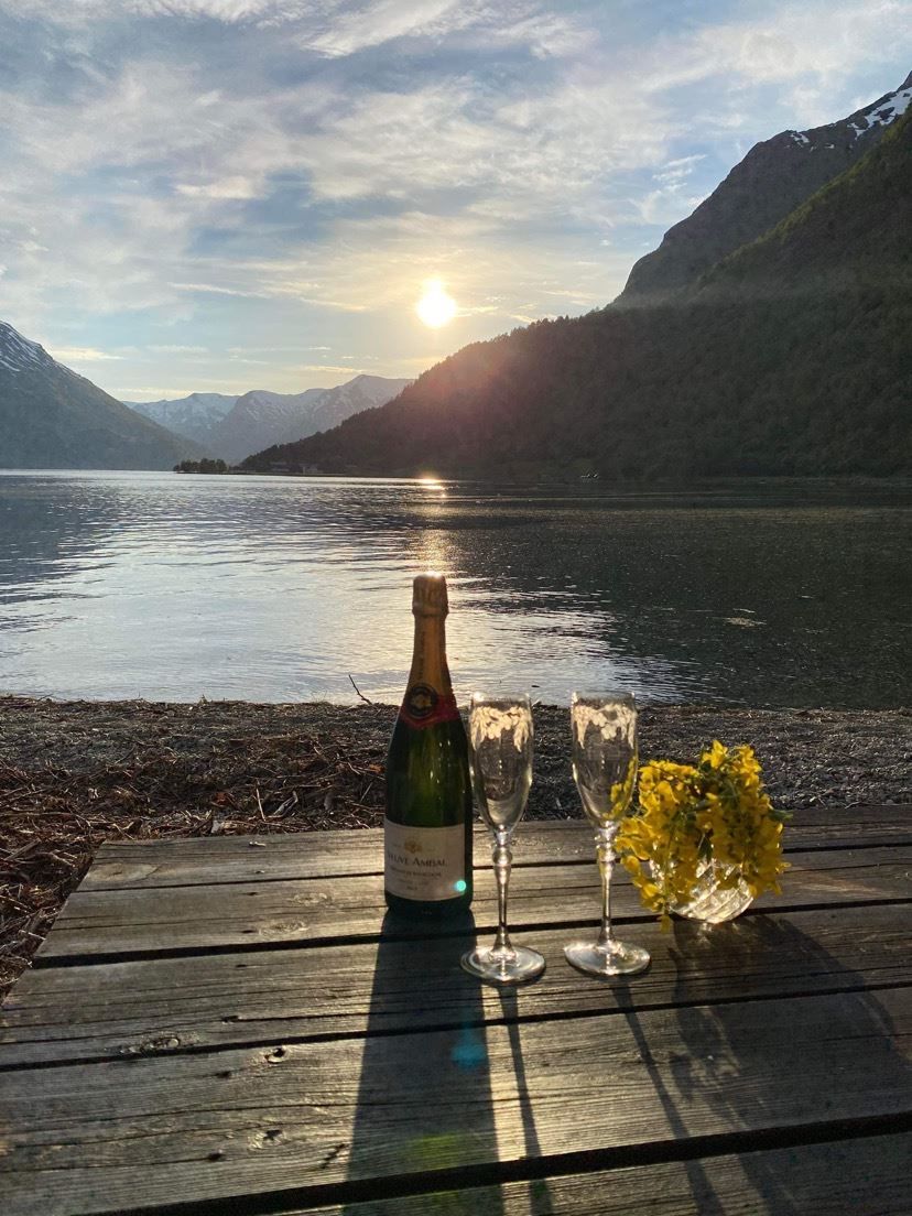 Flaske musserande, to glas og gule blomar på trebord ved stille Oppstrynsvatnet med fjell og låg sol i bakgrunnen på Vollsnes..