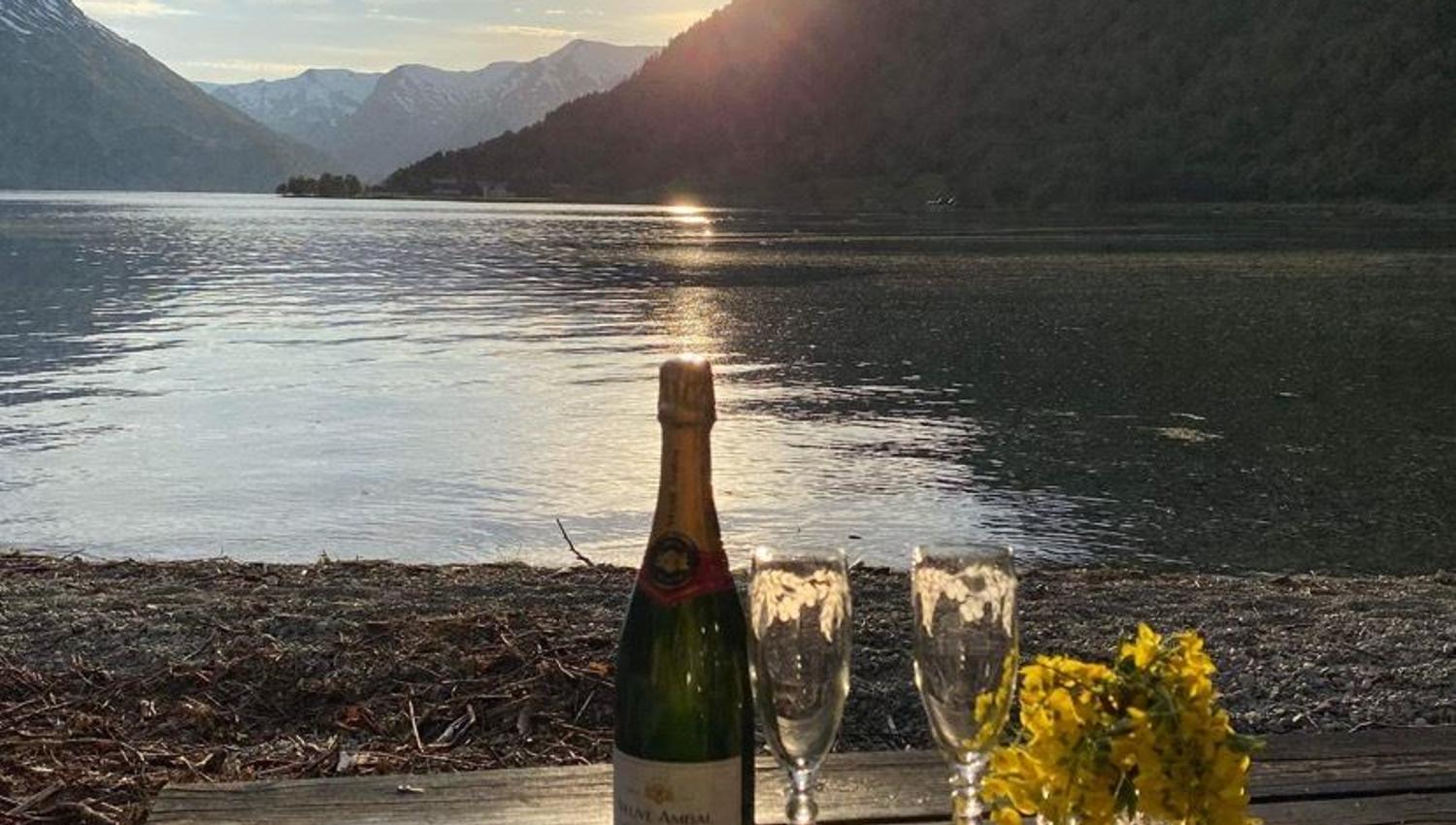 Flaske musserande, to glas og gule blomar på trebord ved stille Oppstrynsvatnet med fjell og låg sol i bakgrunnen på Vollsnes..