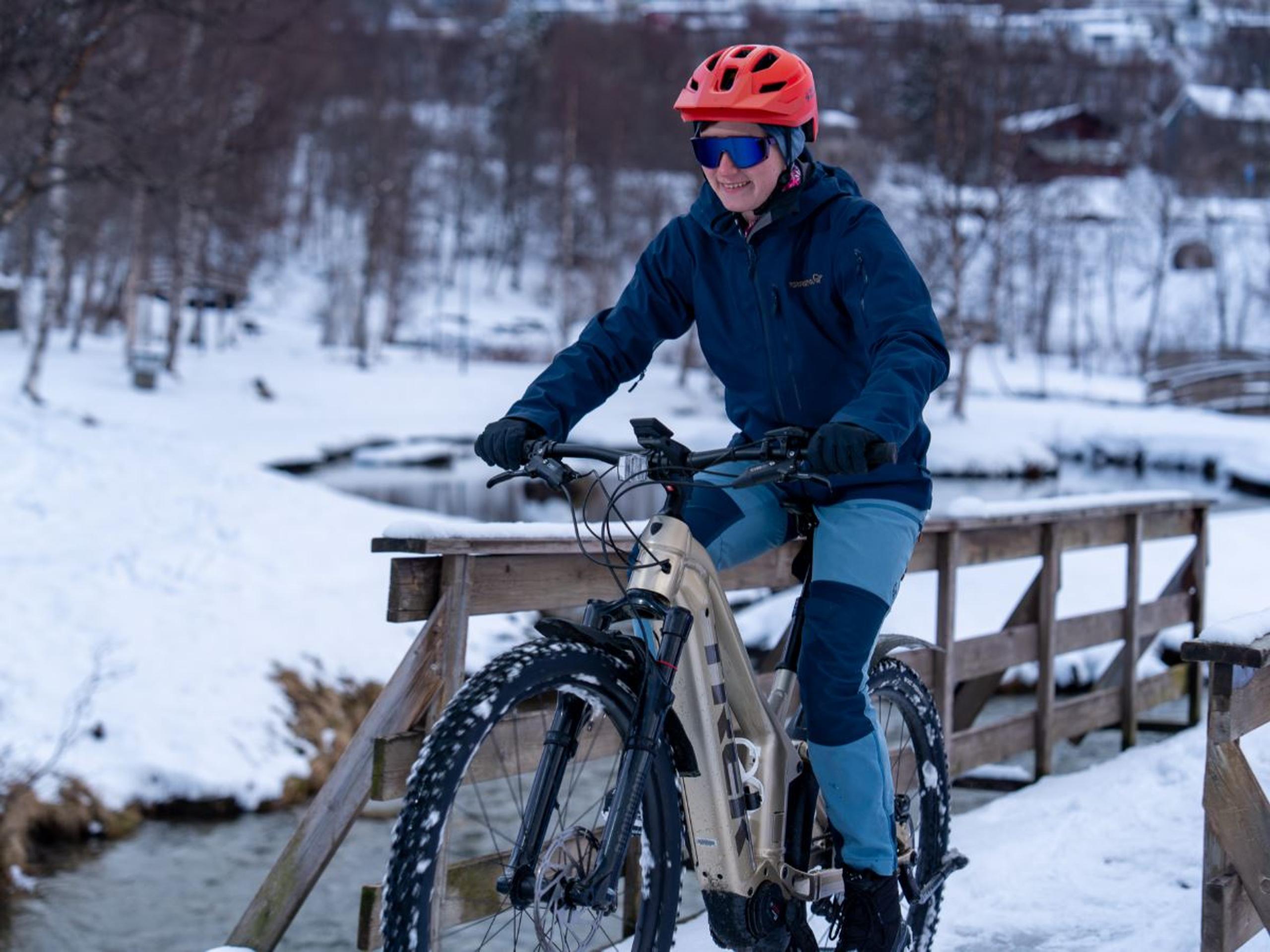 Person wearing a red helmet rides a bike across a wooden bridge over a snowy stream in winter.