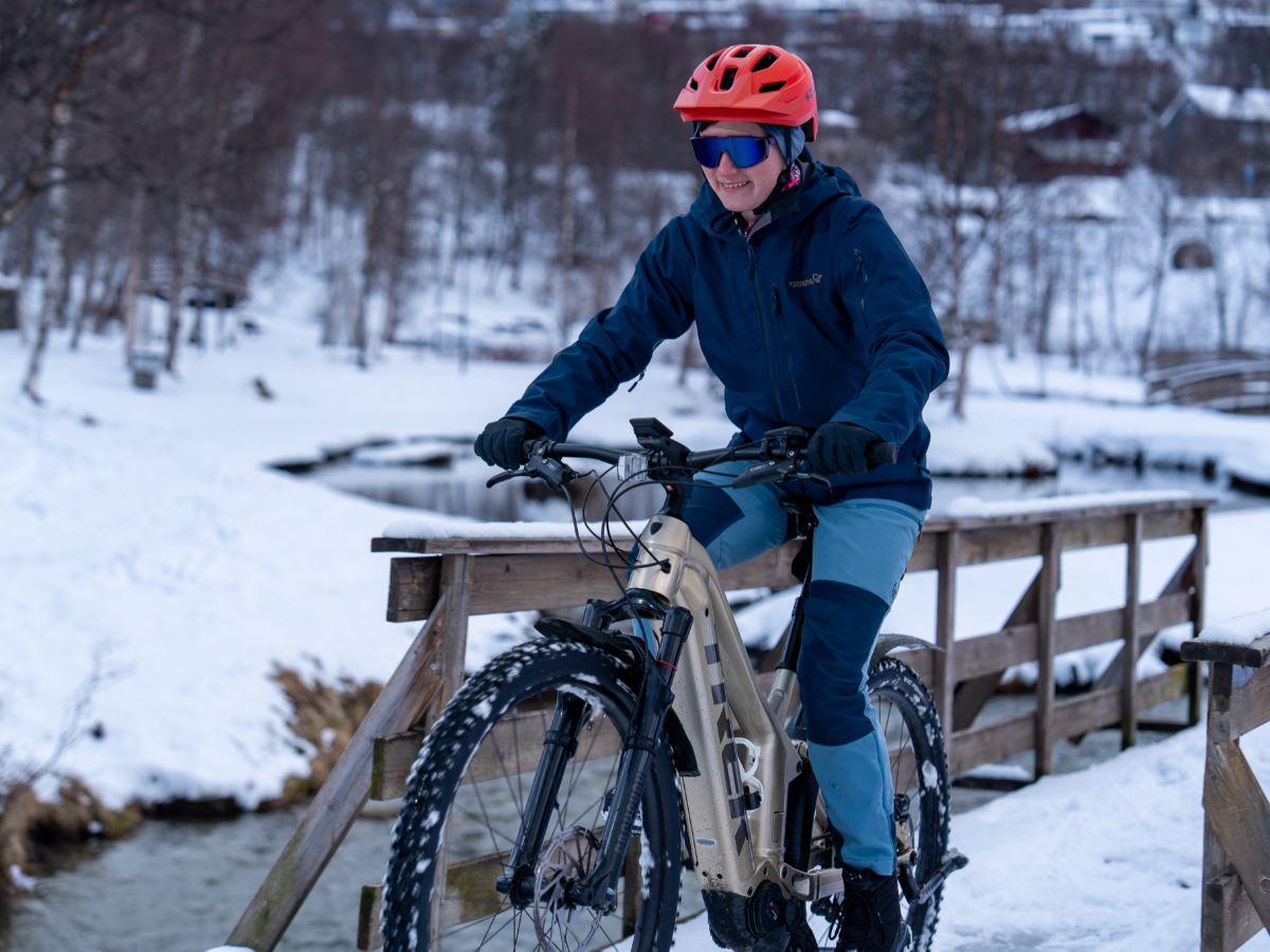 Person wearing a red helmet rides a bike across a wooden bridge over a snowy stream in winter.