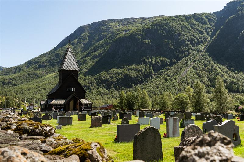 Røldal Stave Church and churchyard in sunlight, surrounded by green hills and steep mountains.