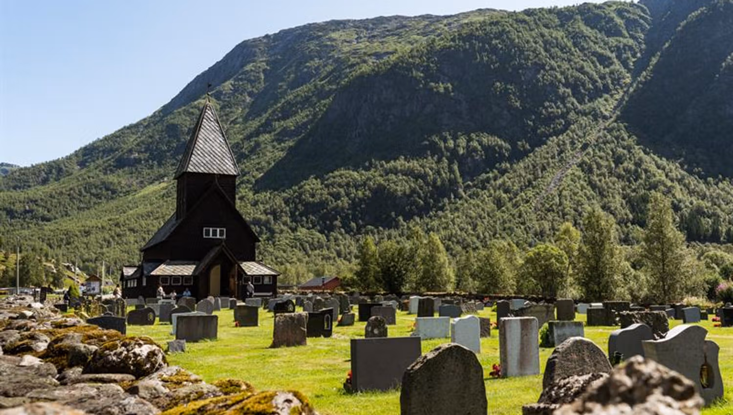 Røldal Stave Church and churchyard in sunlight, surrounded by green hills and steep mountains.