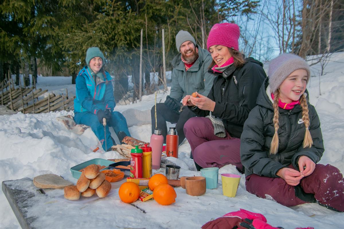 Gruppe som sitter i snøen og spiser niste med termos, kopp og appelsiner.