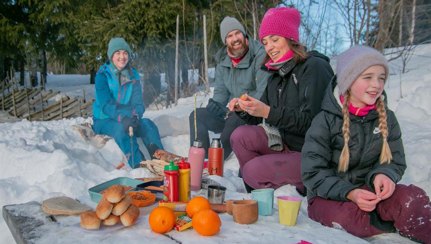 Gruppe som sitter i snøen og spiser niste med termos, kopp og appelsiner.