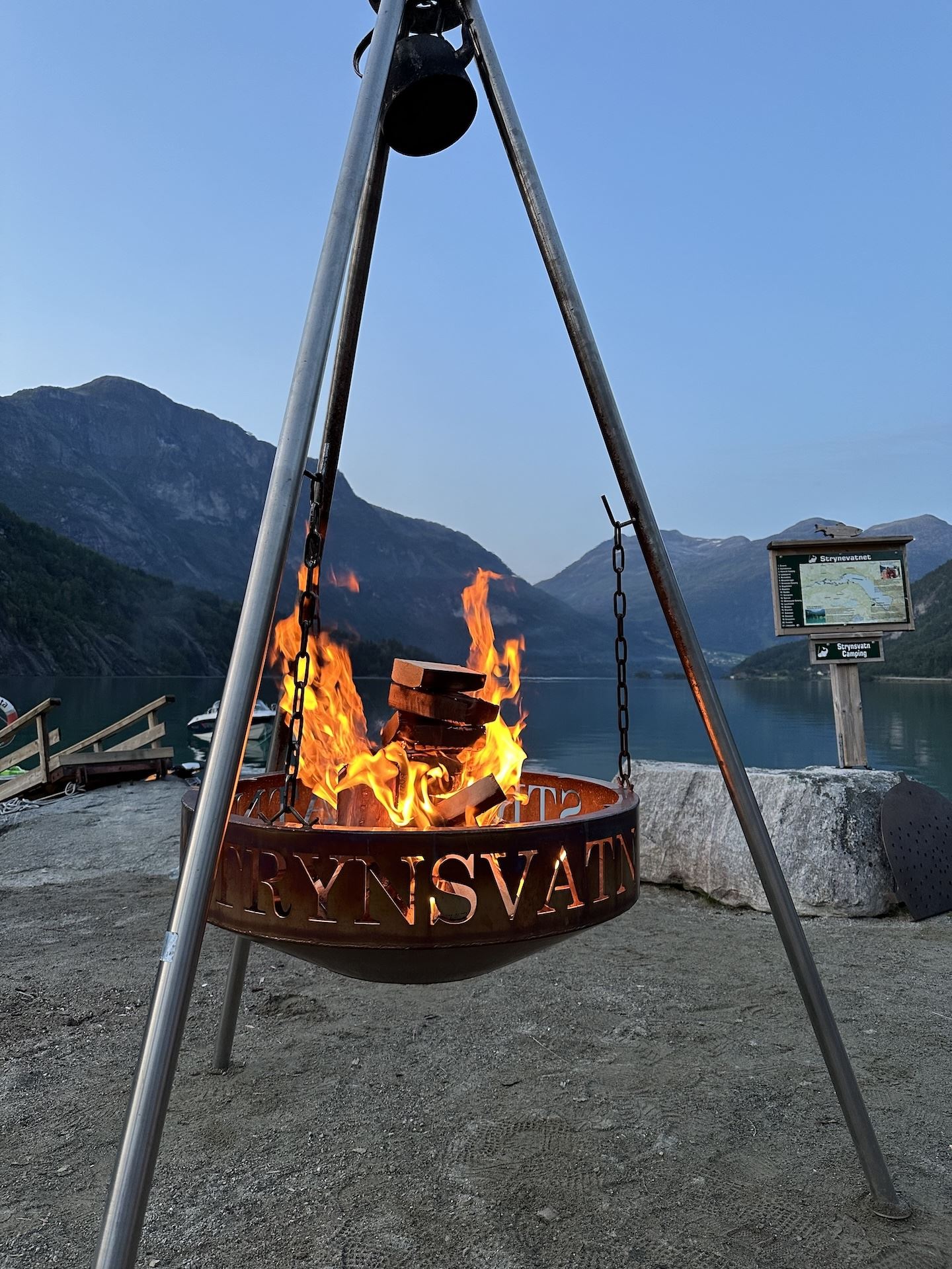 A metal tripod fire pit with burning logs stands by a calm lake, with mountains and a signboard in view..