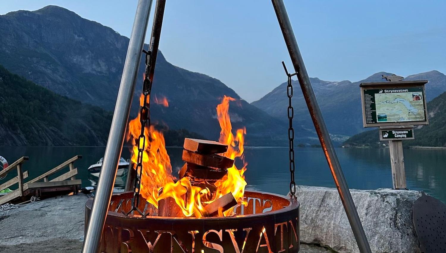 A metal tripod fire pit with burning logs stands by a calm lake, with mountains and a signboard in view..