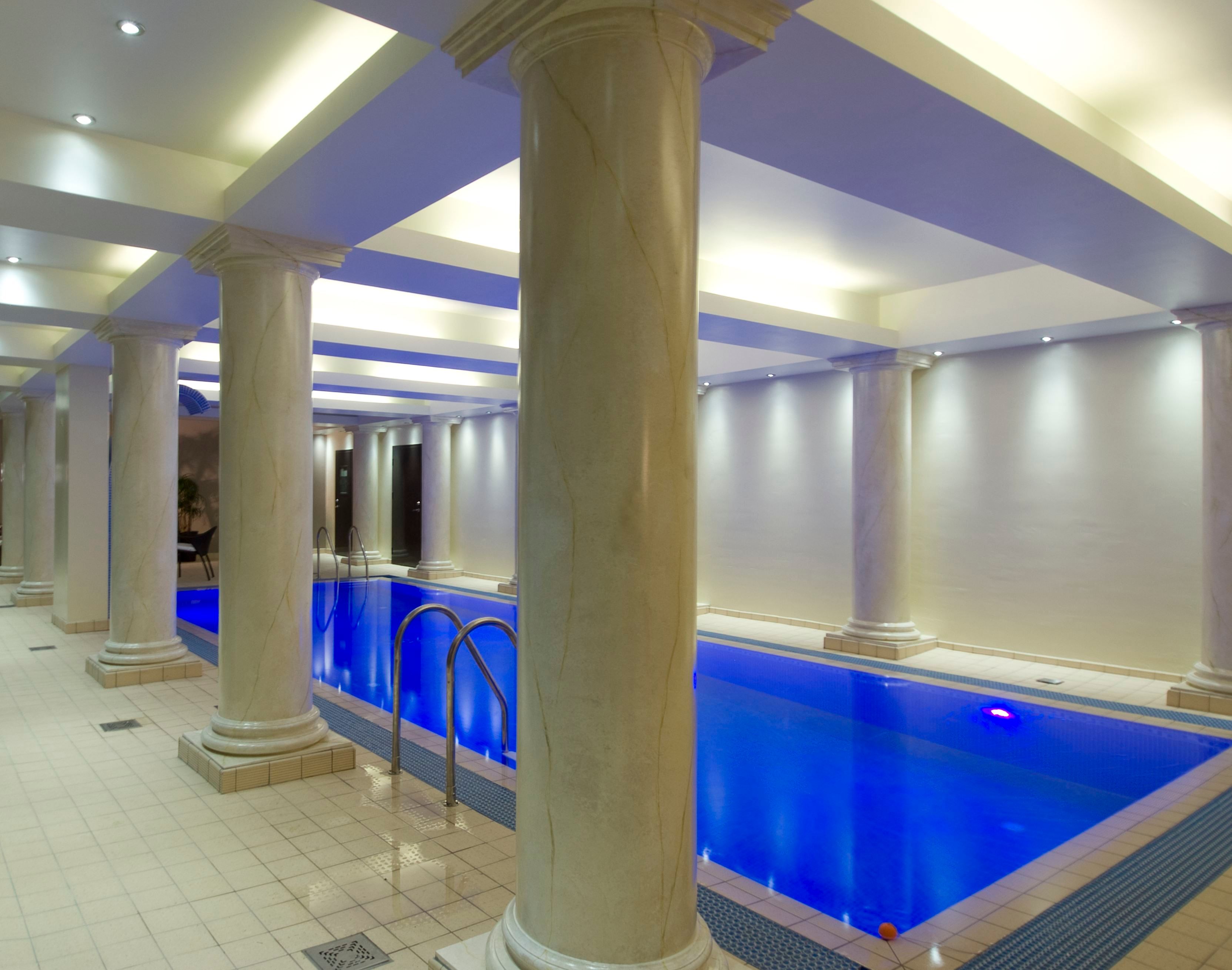 An indoor swimming pool with blue-lit water, marble columns, tiled floor, and recessed ceiling lights.