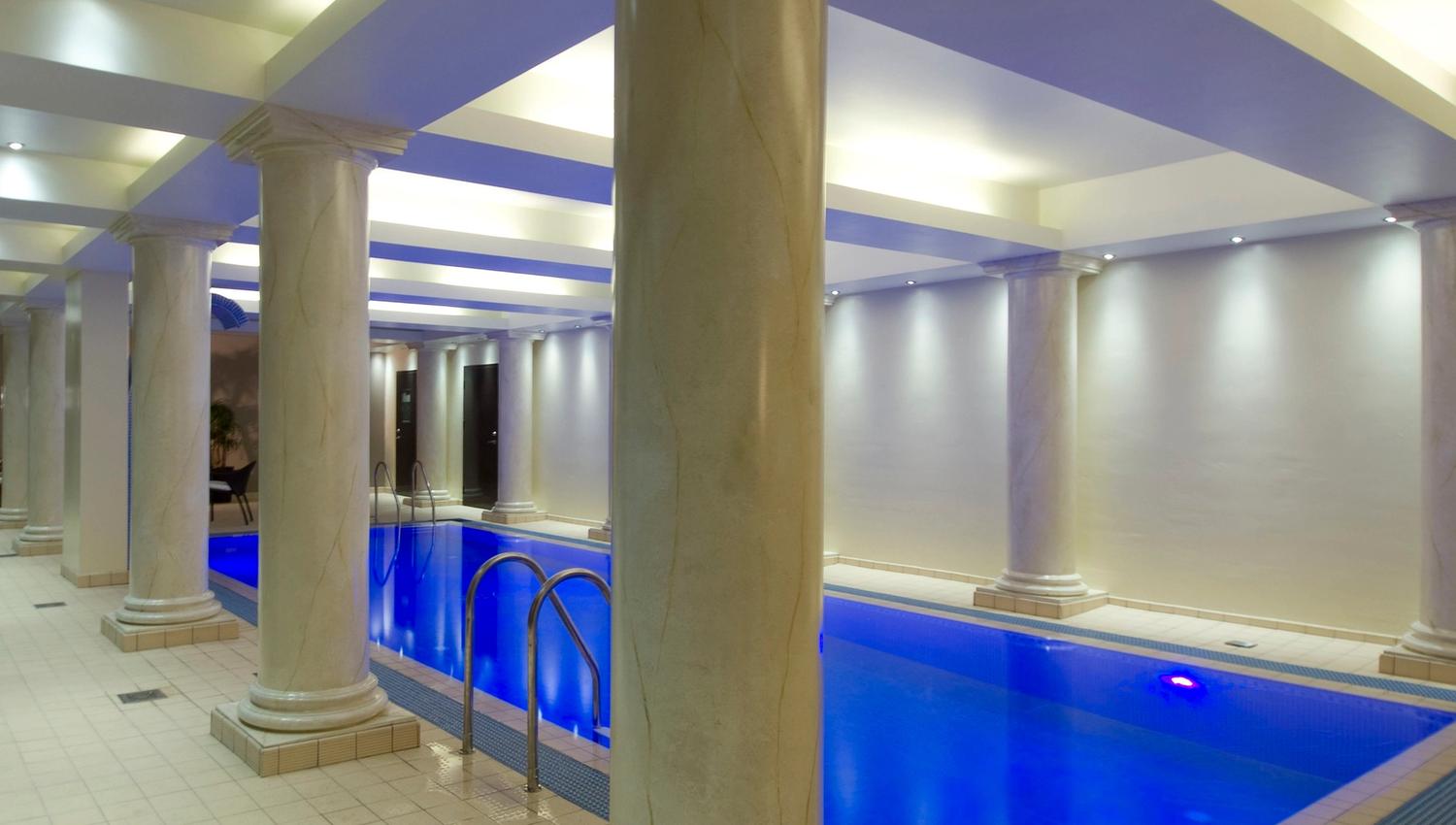 An indoor swimming pool with blue-lit water, marble columns, tiled floor, and recessed ceiling lights.