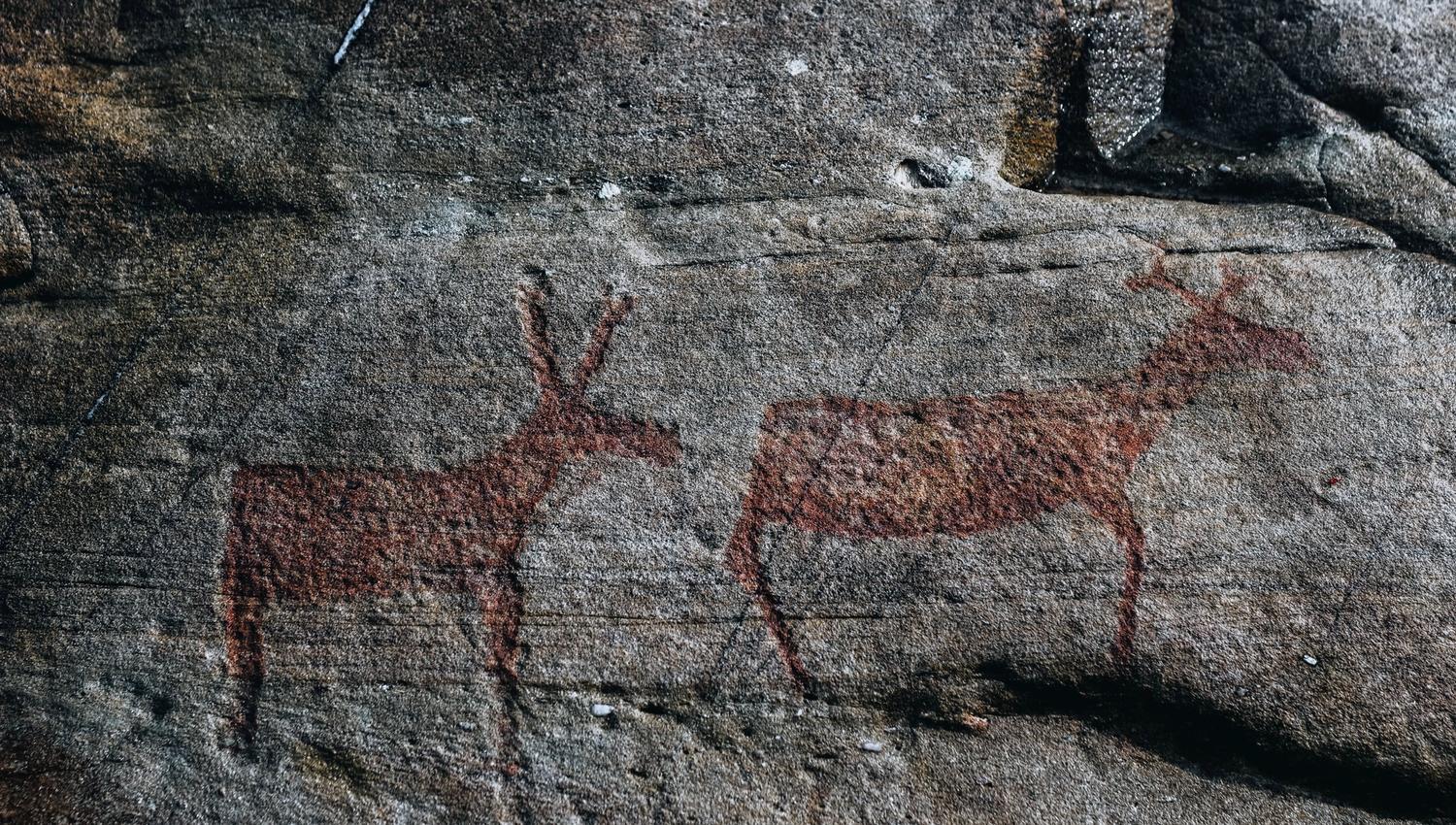Two reddish animal silhouettes painted on a rough gray rock surface, resembling deer in a weathered stone wall.