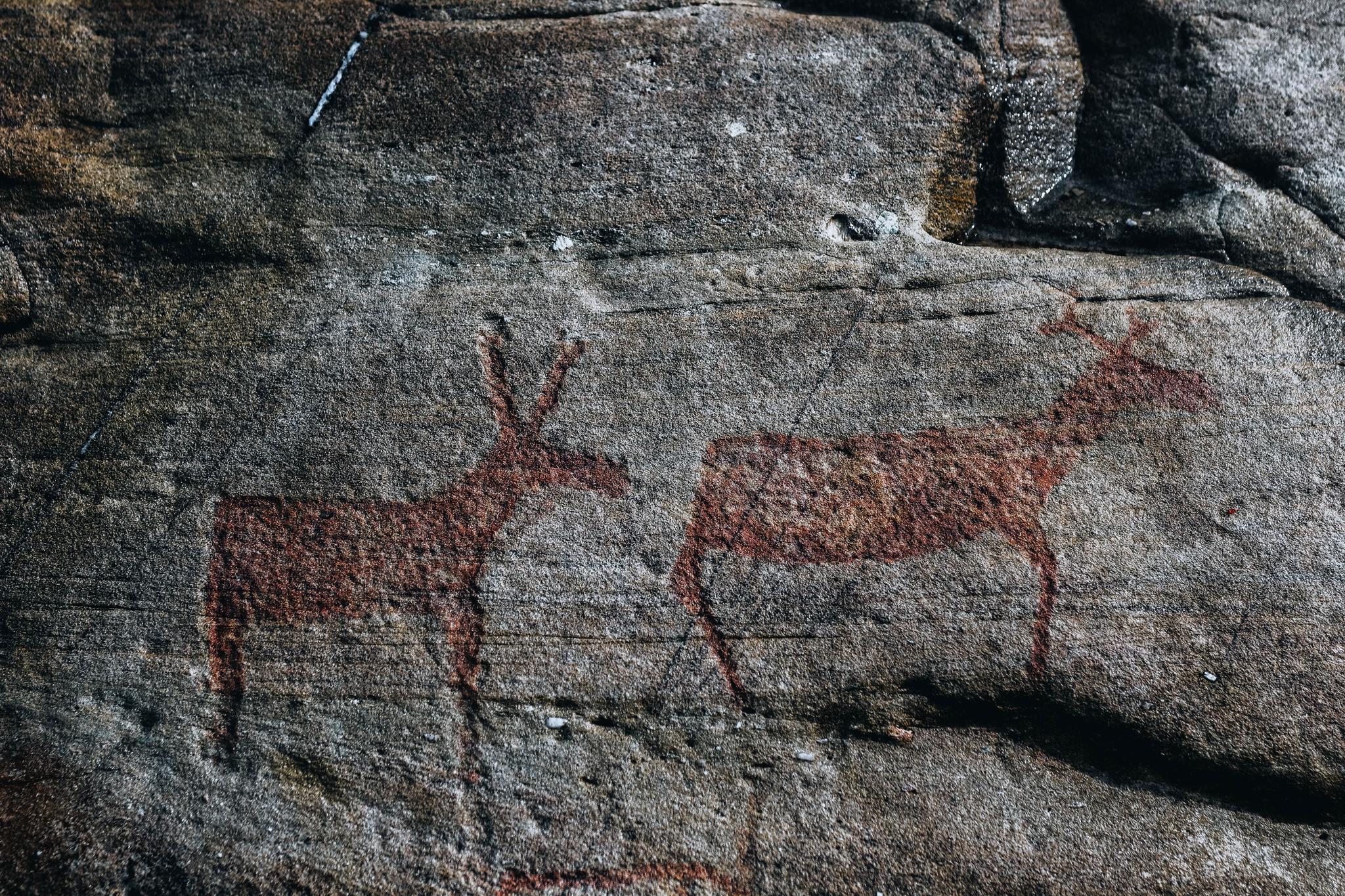 Two reddish animal silhouettes painted on a rough gray rock surface, resembling deer in a weathered stone wall.