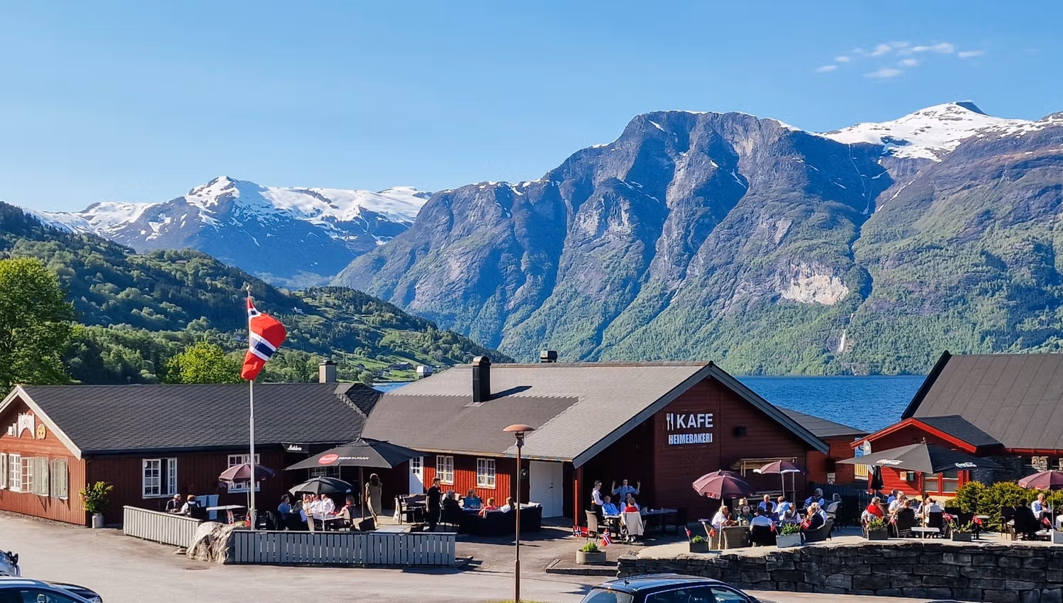 Outdoor café seating beside a lake, with red wooden buildings, a flagpole, and snow-capped mountains under blue sky.
