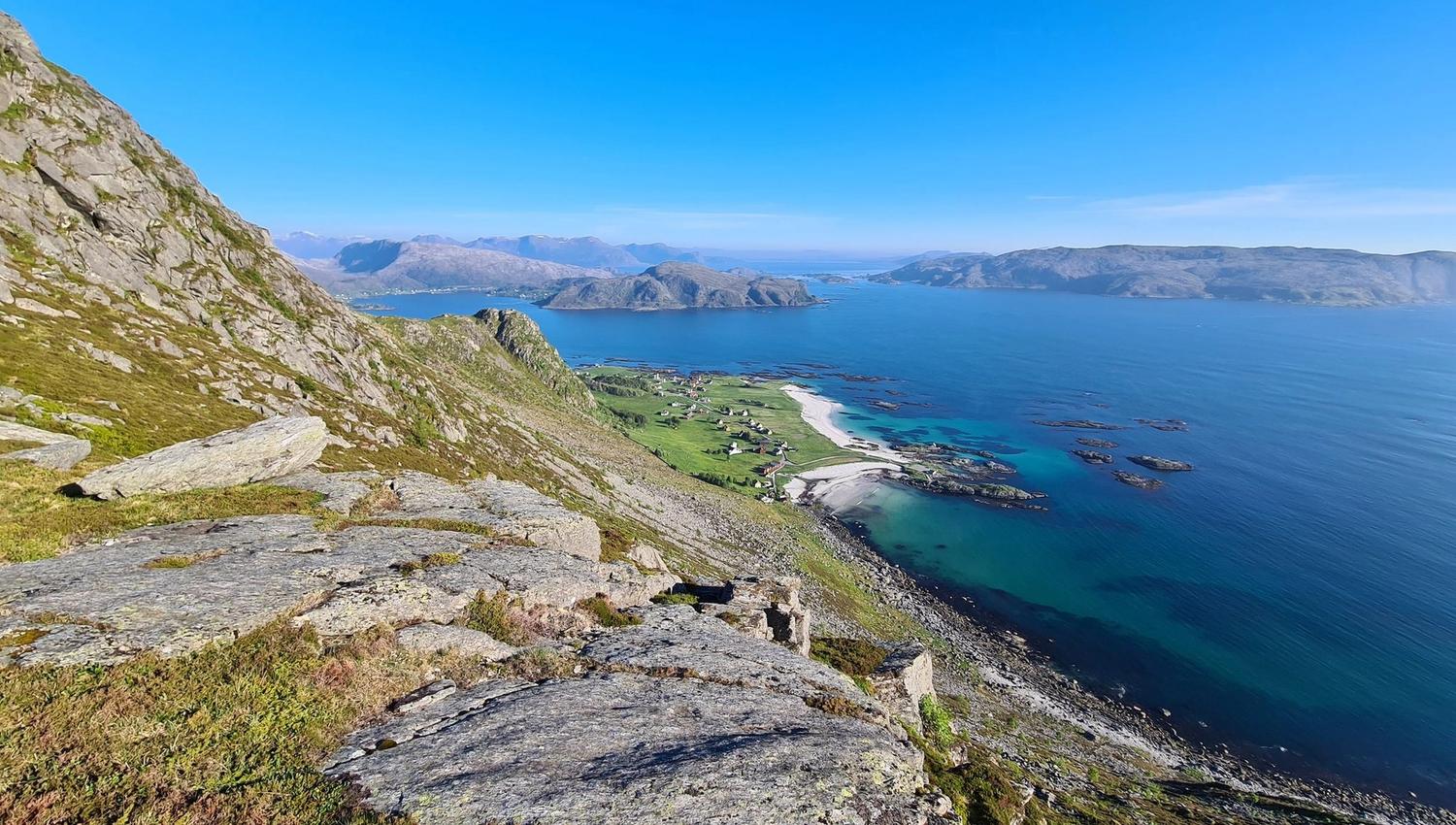 View from a rocky mountainside over green fields, white beaches, and turquoise ocean with distant islands under blue sky.