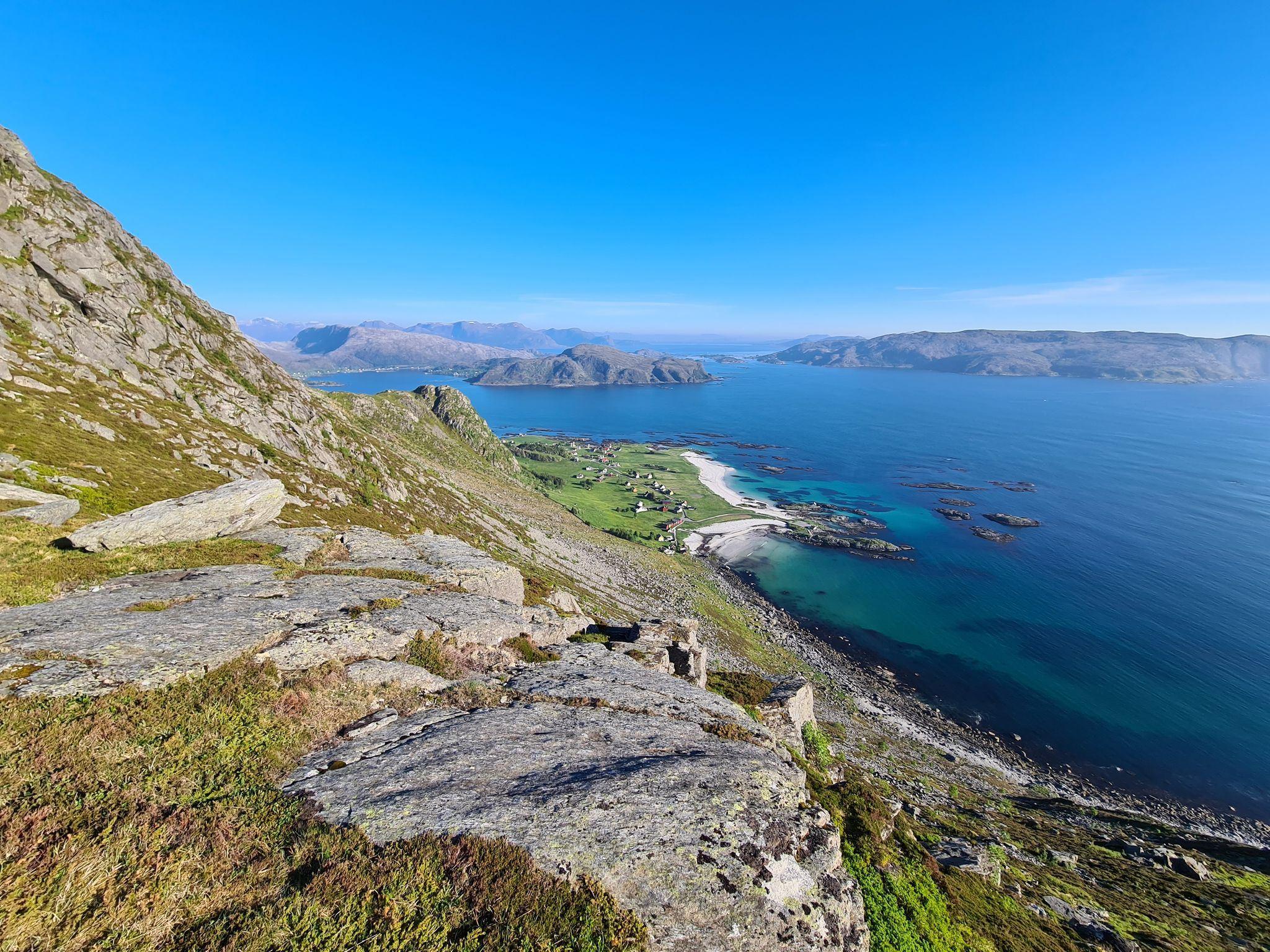 View from a rocky mountainside over green fields, white beaches, and turquoise ocean with distant islands under blue sky.
