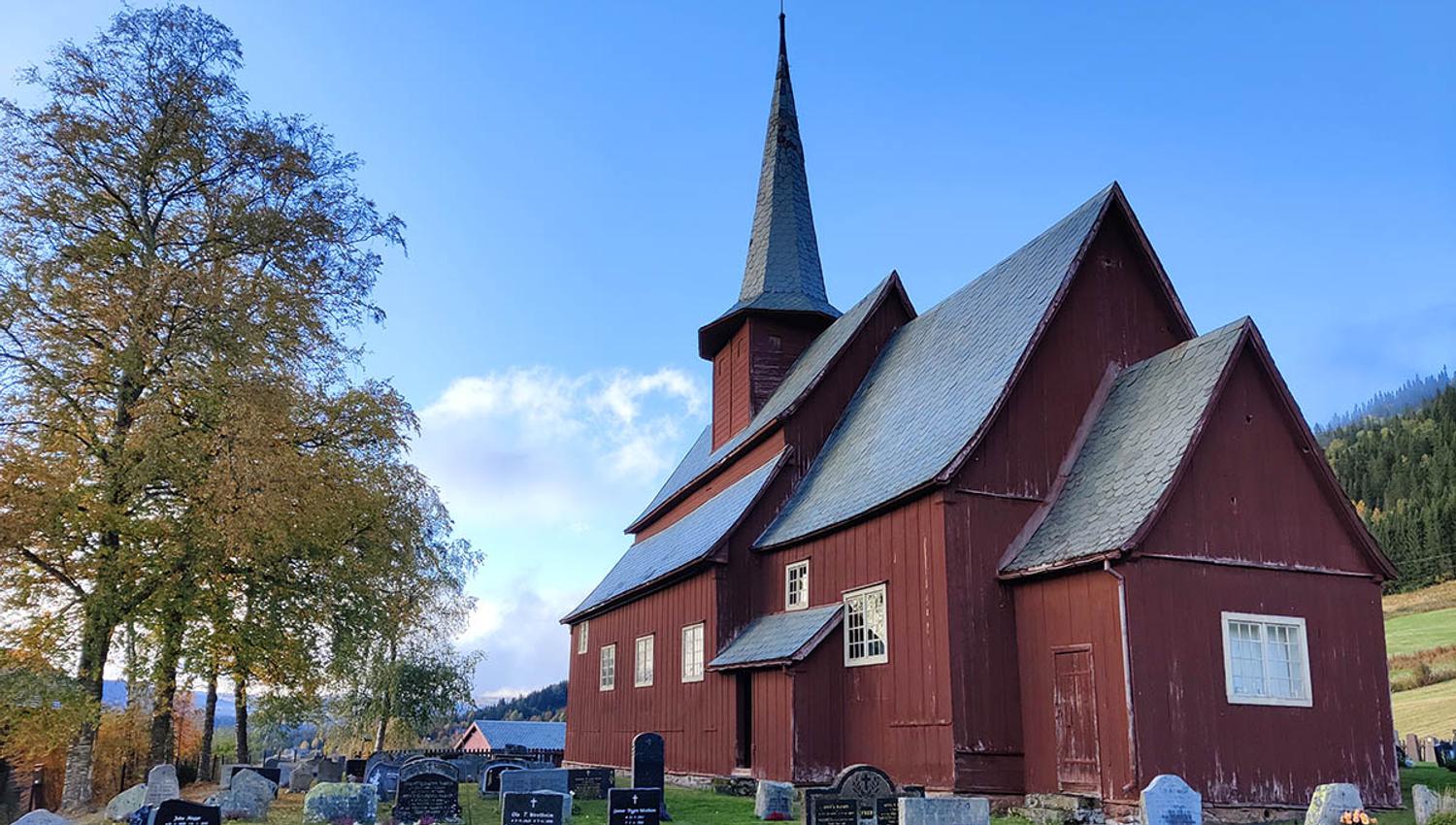 En brunmalt stavkirke i en kirkegard med begynnenede løvfarging tidlig om høsten på bjørketrær like ved.