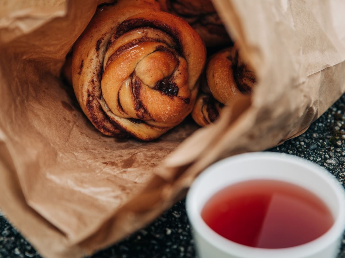 Nærbilde av kanelboller i en brun papirpose, med en hvit kopp rød drikke i forgrunnen..