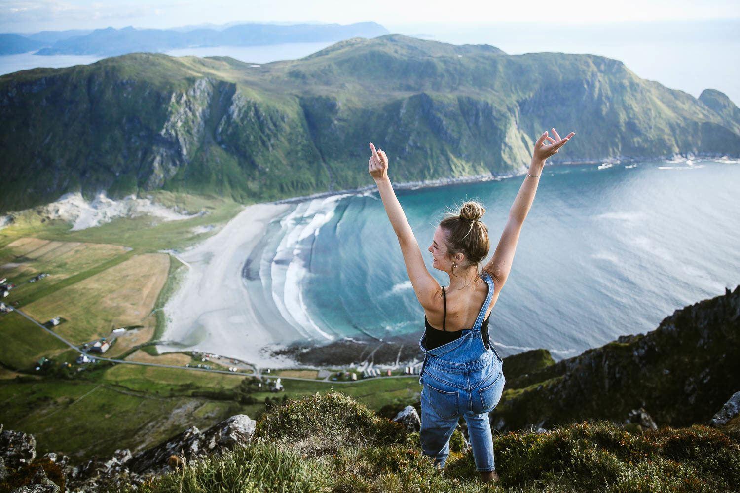 Back view of a woman on a cliff with arms raised, overlooking a curved beach, blue ocean, and green mountains.