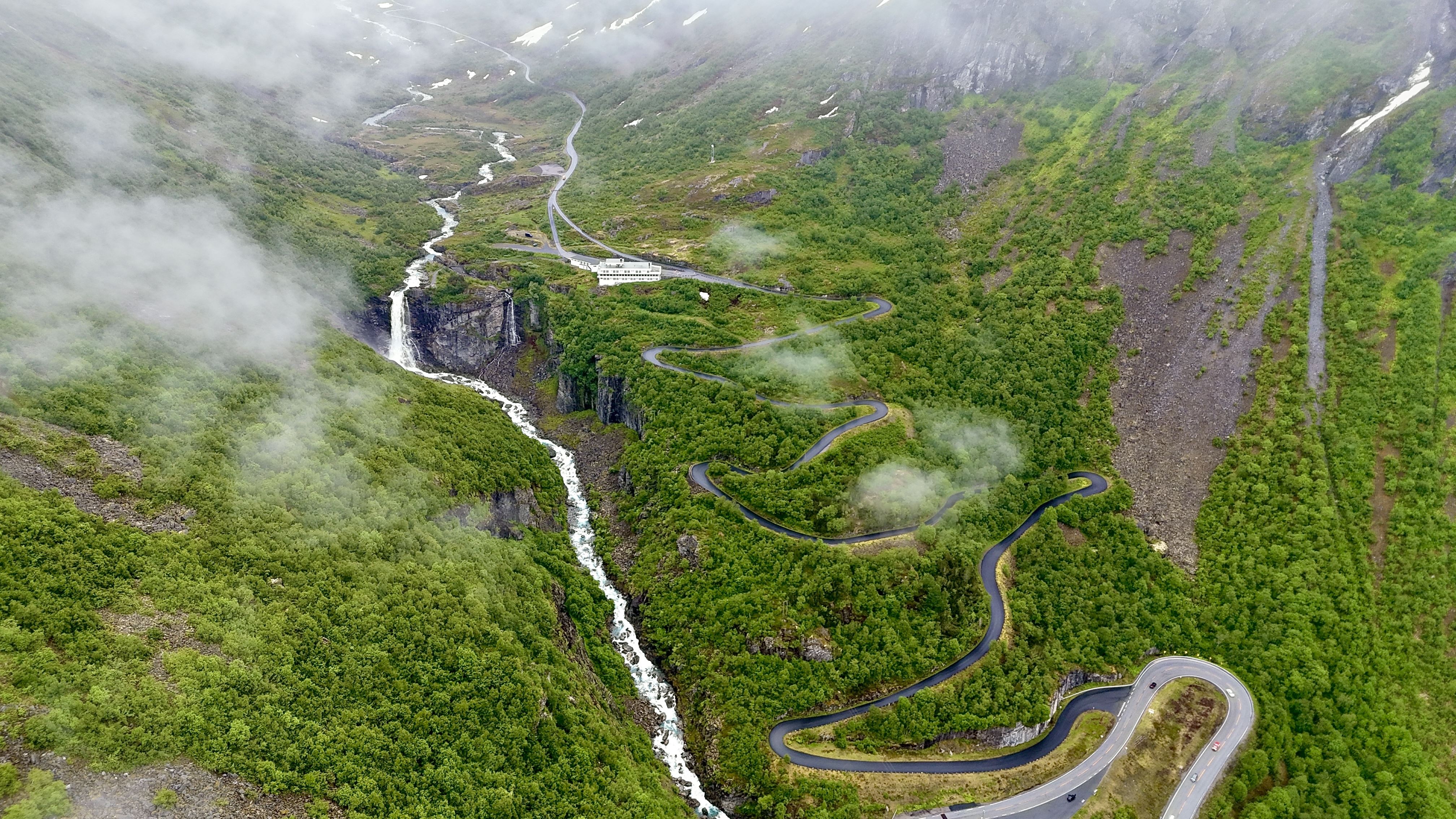 Aerial view of a winding road through a green mountain valley with a waterfall and low clouds drifting over slopes..