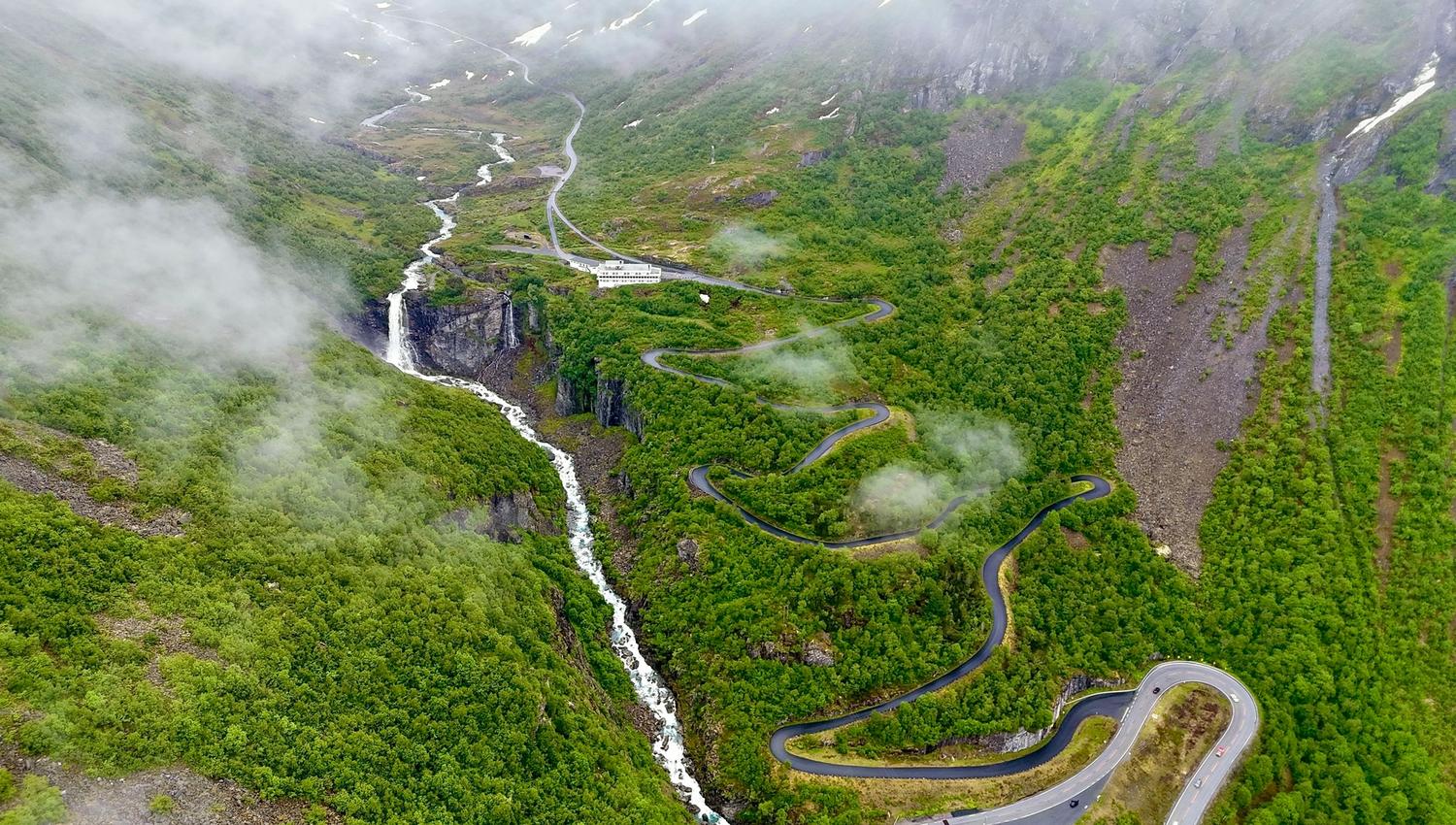 Aerial view of a winding road through a green mountain valley with a waterfall and low clouds drifting over slopes..