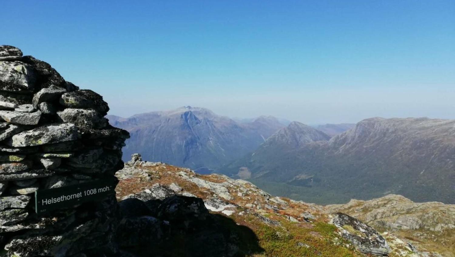 Stone cairn on a rocky ridge with a small sign, overlooking distant mountain peaks and a broad valley under blue sky..