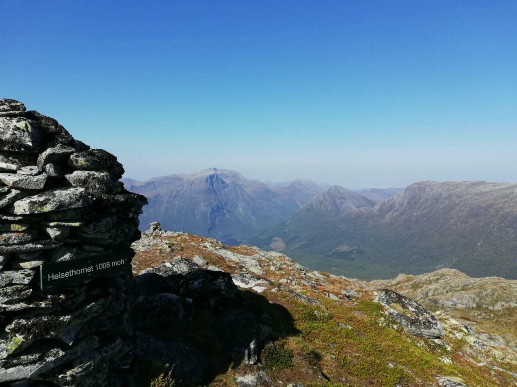 Stone cairn on a rocky ridge with a small sign, overlooking distant mountain peaks and a broad valley under blue sky..