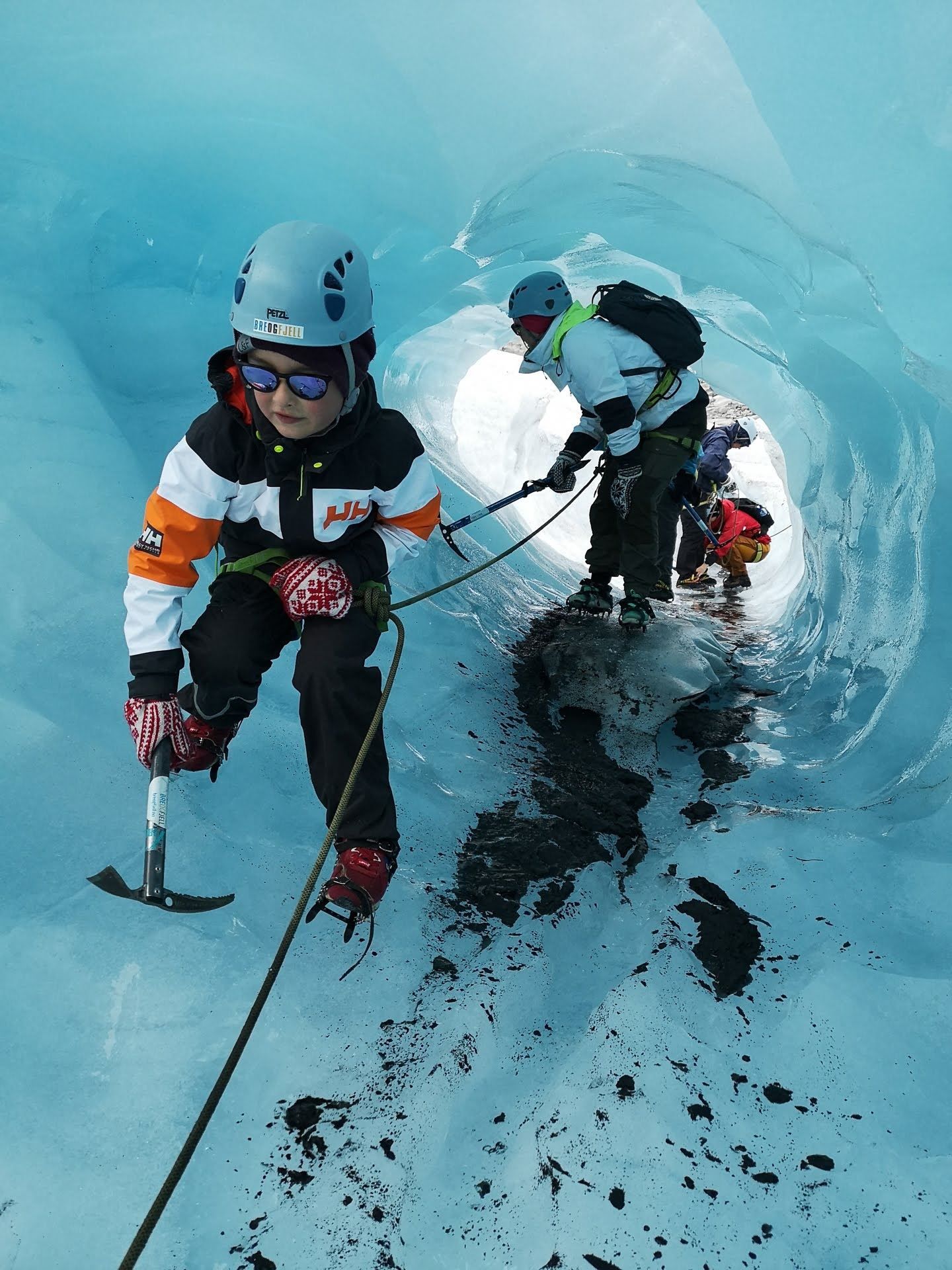 Roped ice climbers wearing helmets use ice axes and crampons while moving through a blue glacier tunnel..