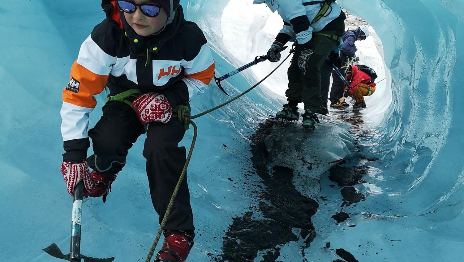 Roped ice climbers wearing helmets use ice axes and crampons while moving through a blue glacier tunnel..