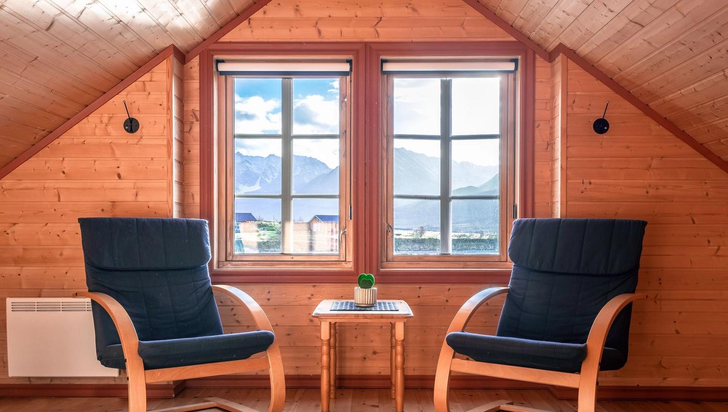 Attic lounge with two armchairs, a wooden table, and views of mountains and houses through large windows