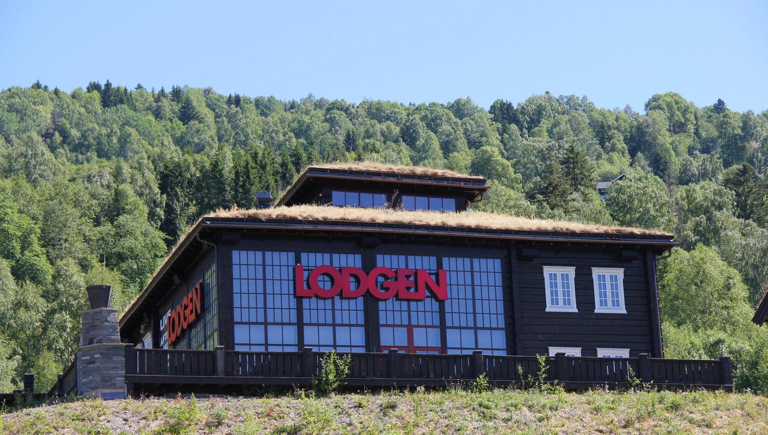 Mørk trebygning med store vinduer og rød «LODGEN»-logo, gress på taket og skogkledd åsside bak..