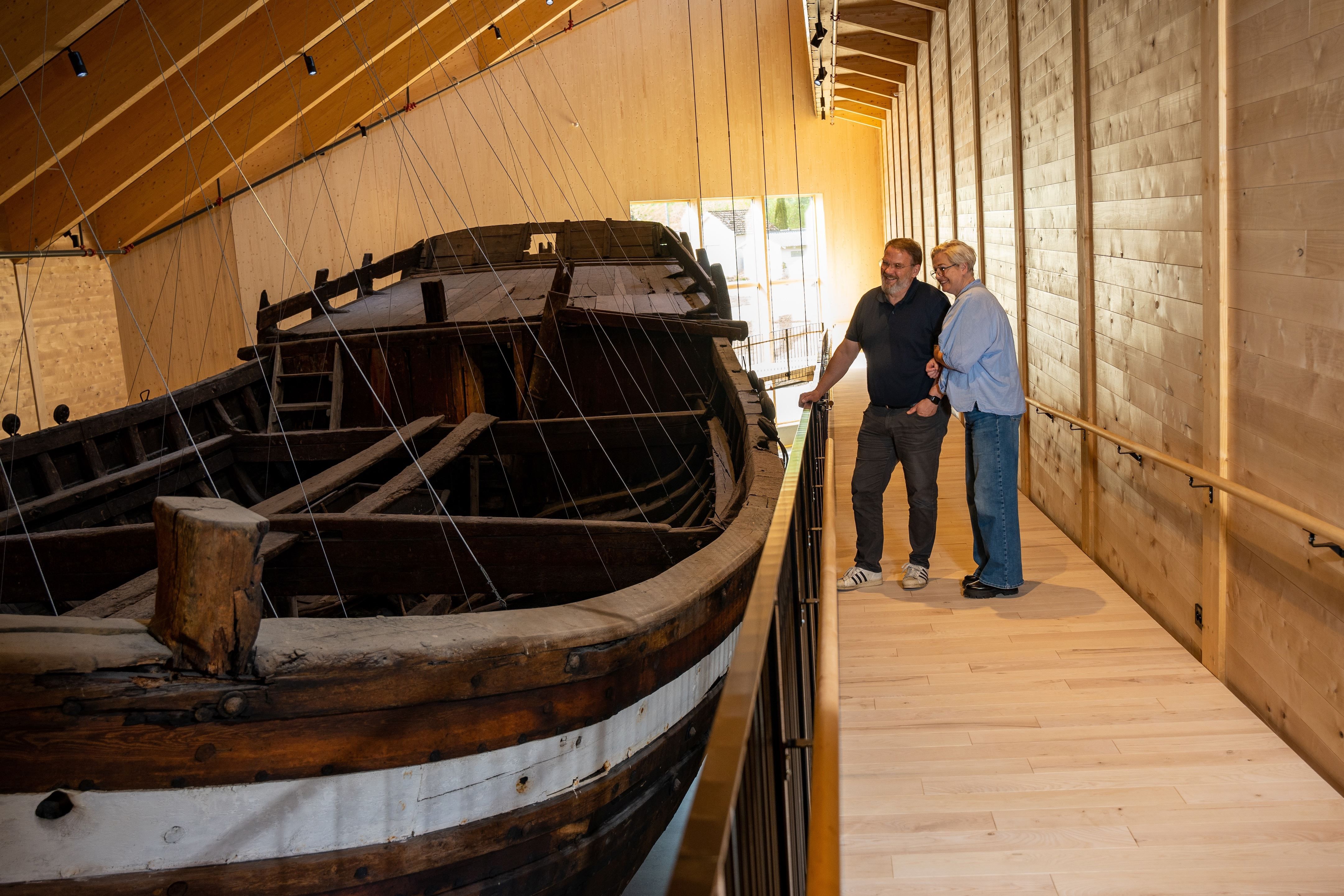 Two visitors stand on a wooden walkway beside a large old wooden boat, Holvikejekta, displayed indoors under a sloped roof..