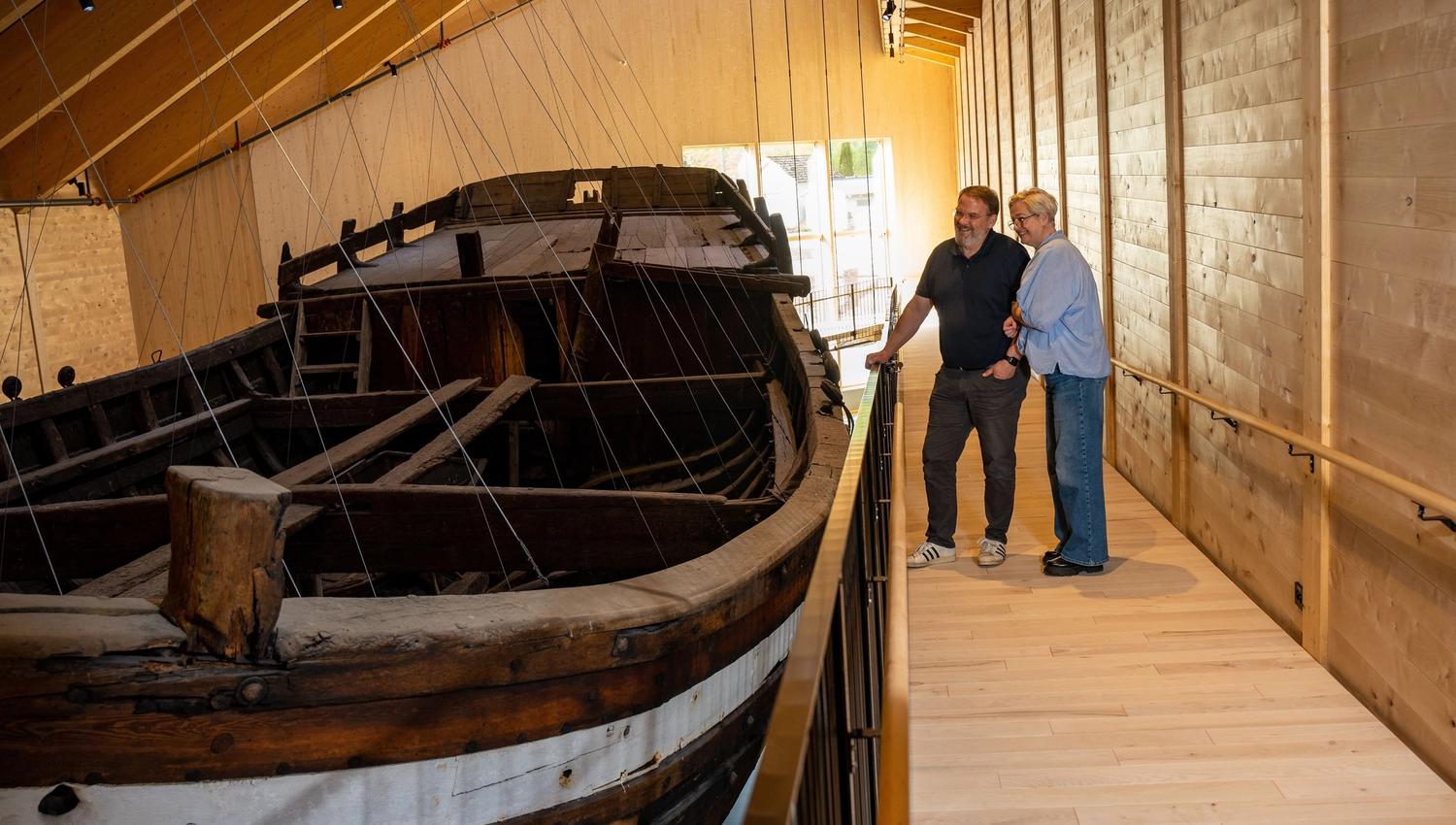 Two visitors stand on a wooden walkway beside a large old wooden boat, Holvikejekta, displayed indoors under a sloped roof..