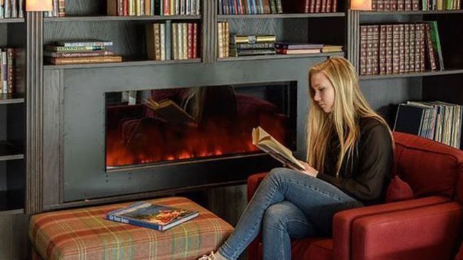 Person reading a book in a red armchair in front of an electric fireplace and bookshelves