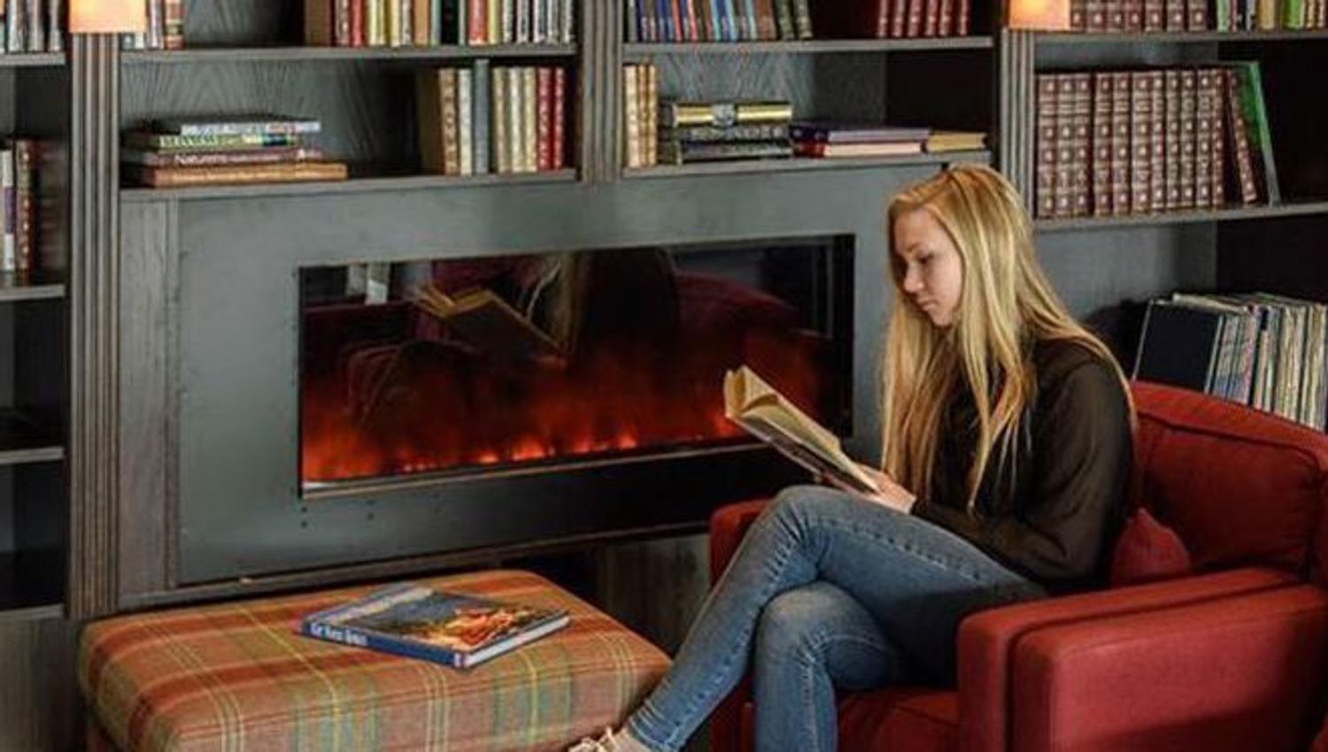 Person reading a book in a red armchair in front of an electric fireplace and bookshelves
