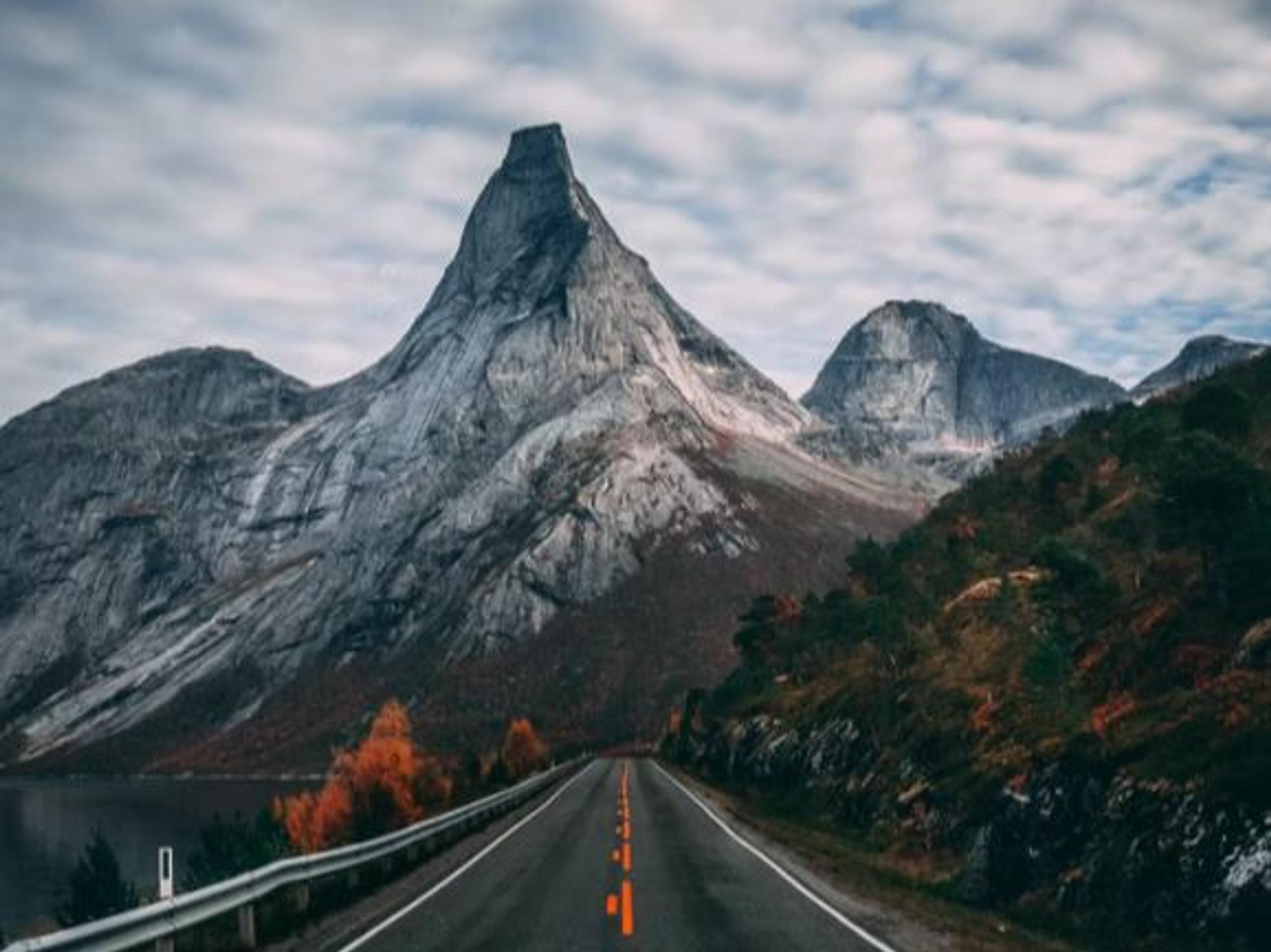 En tom asfaltvei leder inn i en dal mot et spisst, grått fjellmassiv under overskyet himmel..