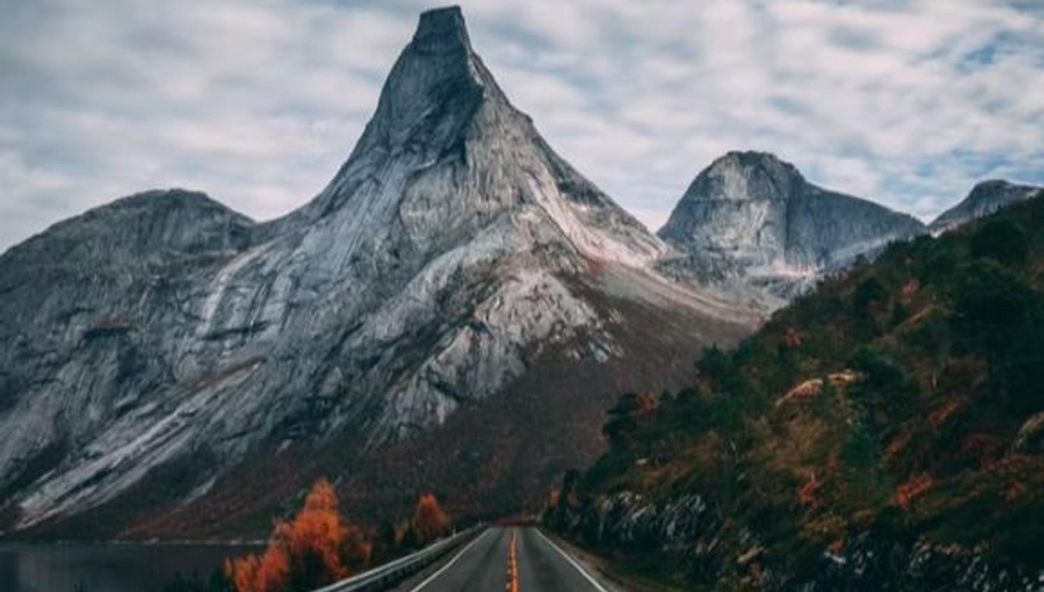En tom asfaltvei leder inn i en dal mot et spisst, grått fjellmassiv under overskyet himmel..