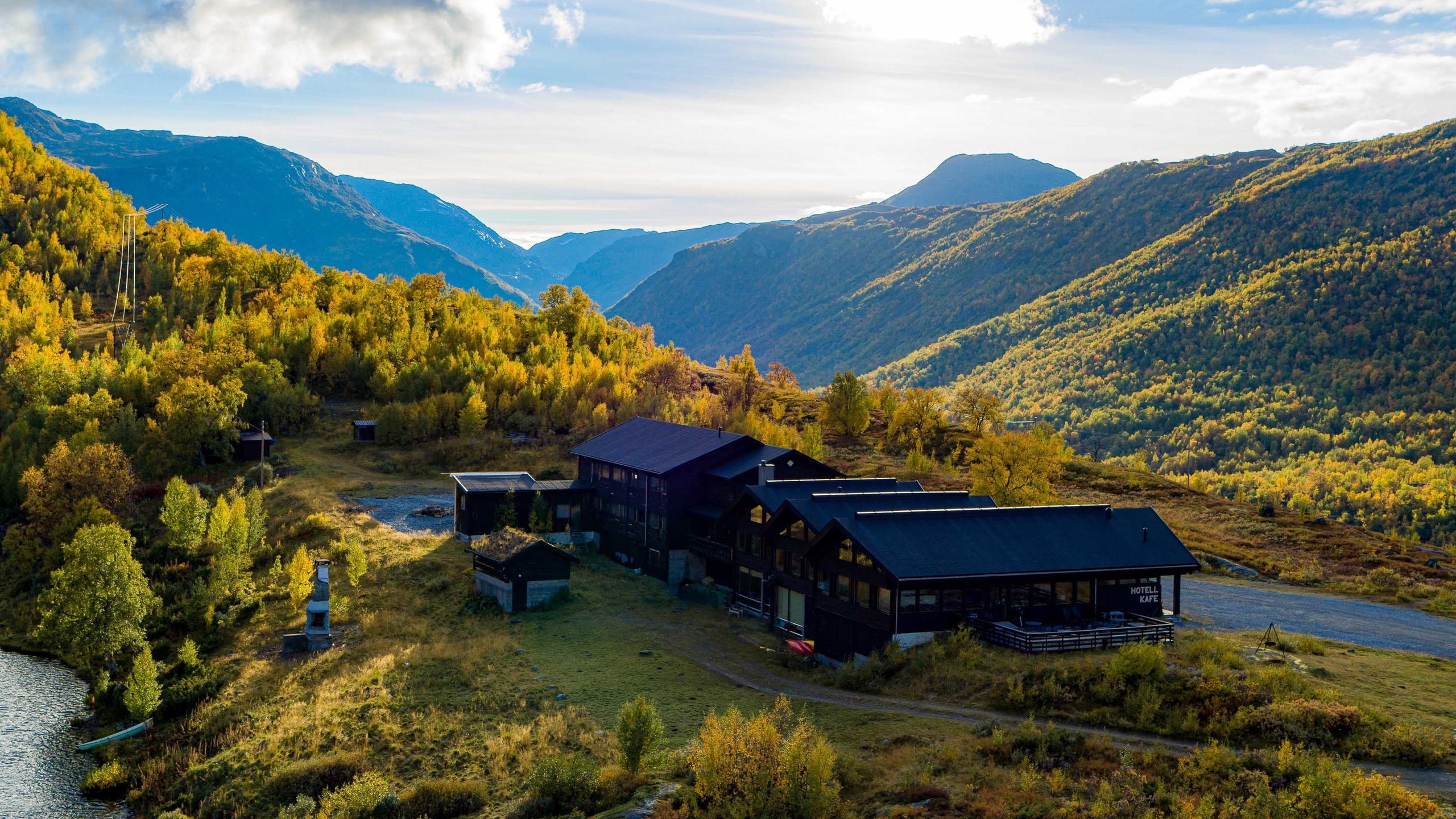 Mørk fjellhytte ved ei fjellside med haustfargar, skog og dalar, med fjell og skyer i bakgrunnen..
