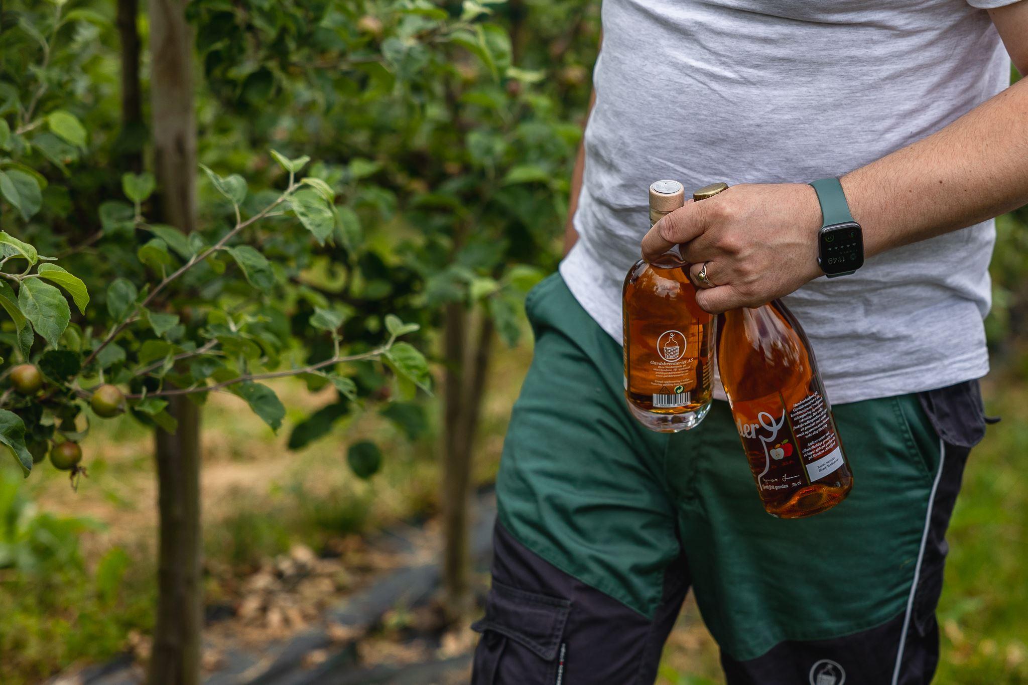 Close-up of a person holding two glass bottles of amber liquid beside leafy fruit trees in an orchard.