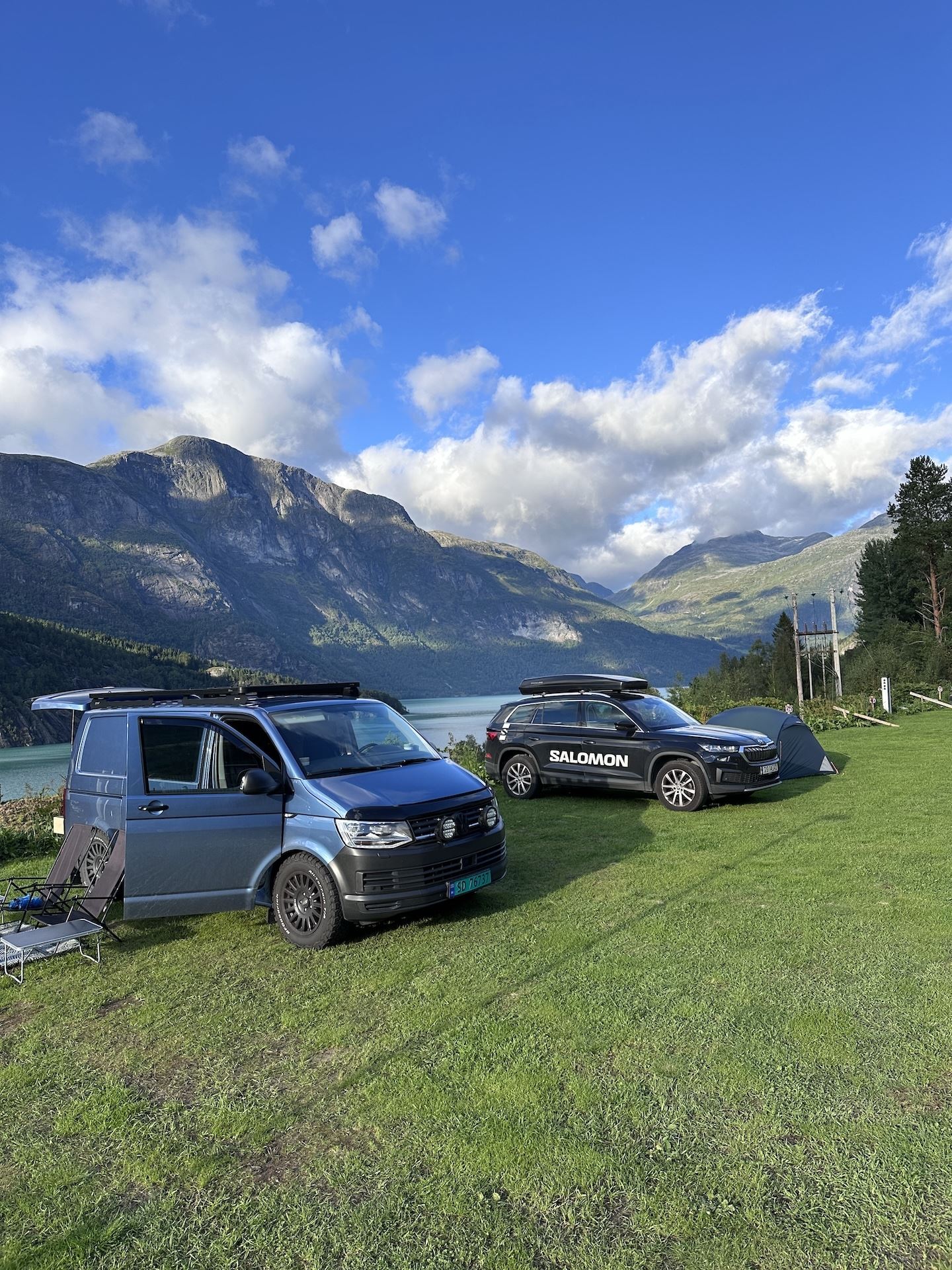 Two camper vehicles and a small tent on a grassy lakeside with rugged mountains and clouds under blue sky..