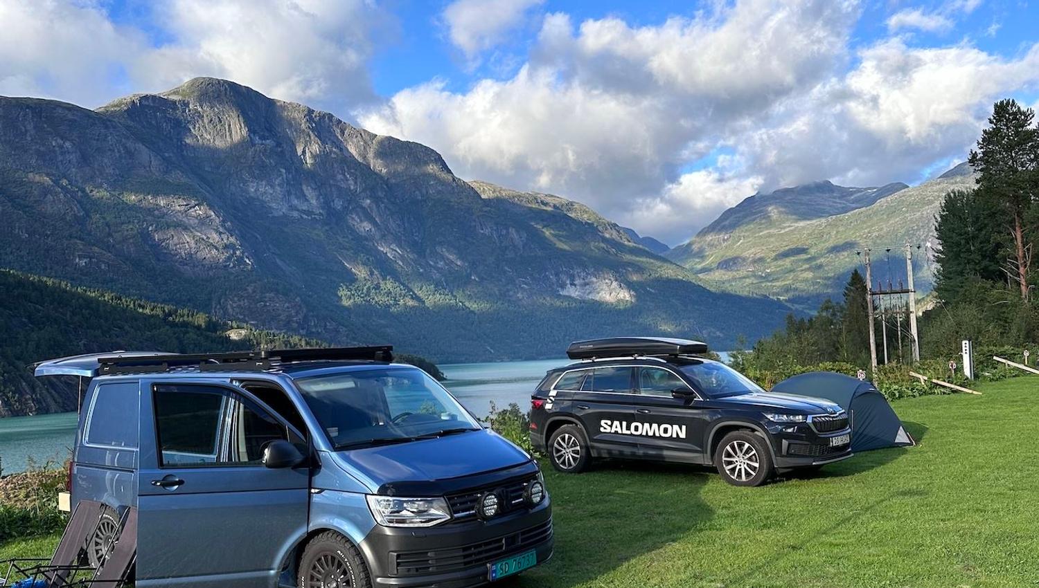 Two camper vehicles and a small tent on a grassy lakeside with rugged mountains and clouds under blue sky..